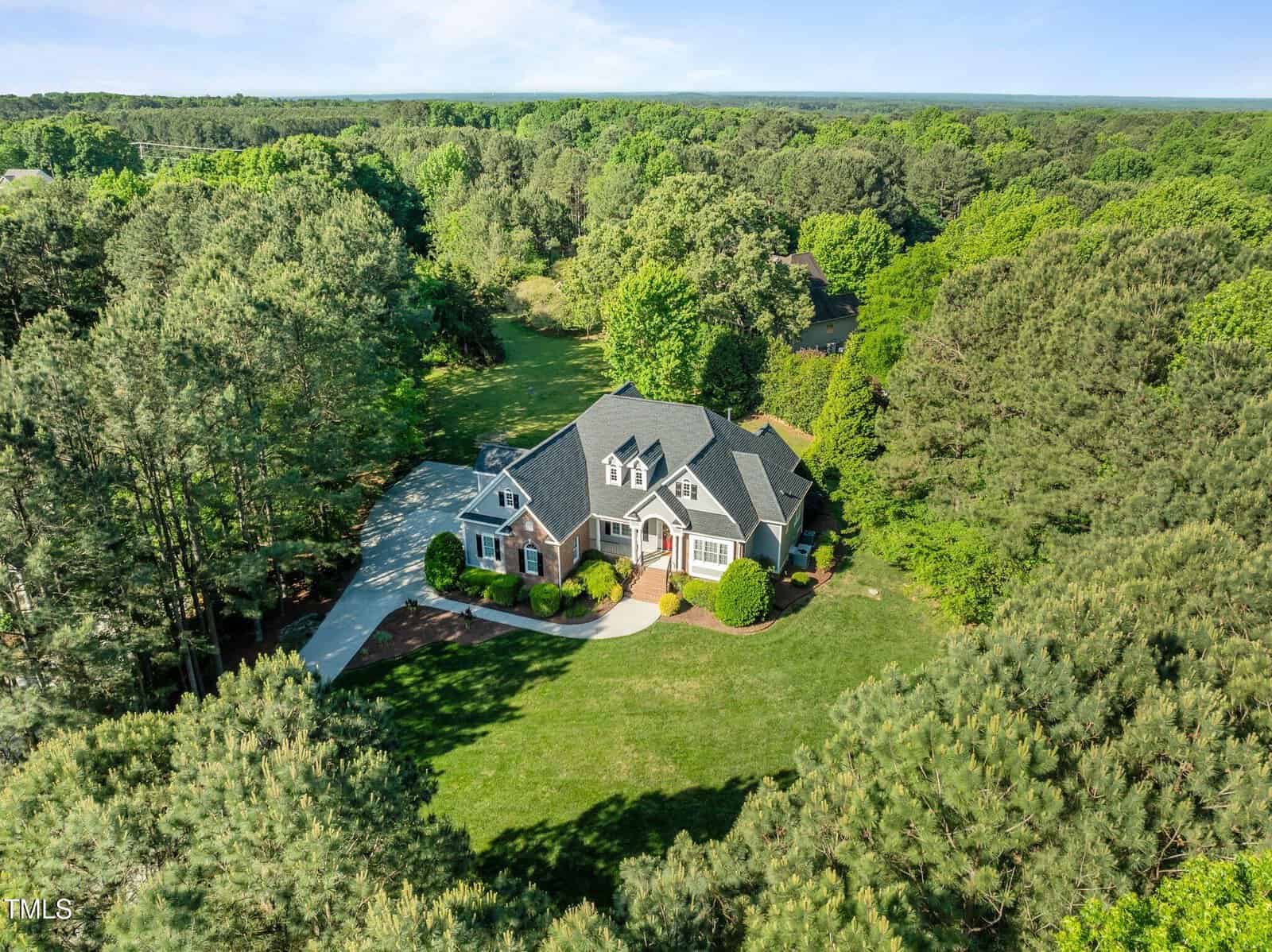 A large two-story house with a well-maintained lawn and driveway is surrounded by dense trees and greenery, set in a secluded, wooded area under a clear sky.