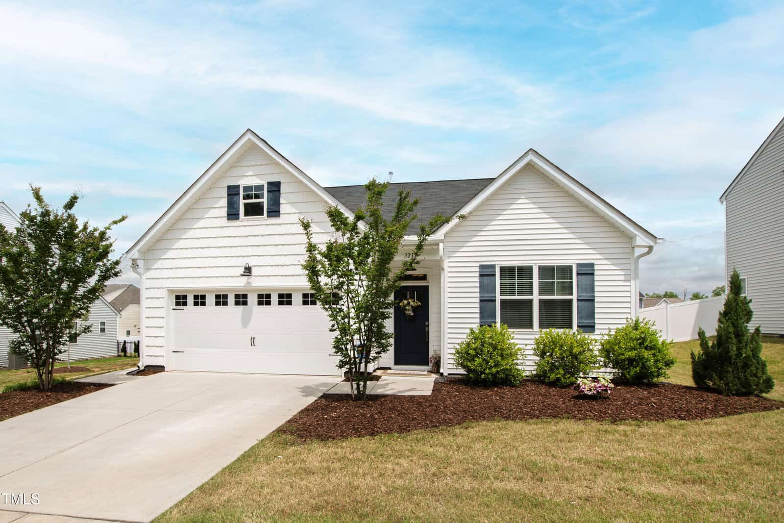 A single-story white house with black shutters, a two-car garage, a dark front door, small trees, and bushes in the front yard. The house sits on a concrete driveway, under a partly cloudy sky.