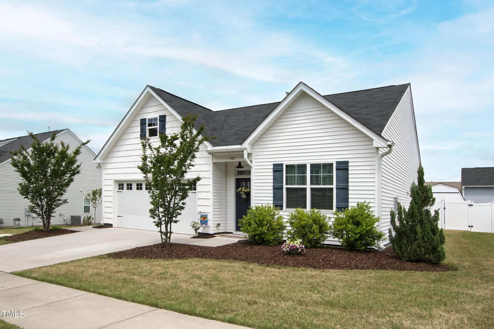 A single-story white house with black shutters, a front lawn, shrubs, and a driveway leading to a two-car garage, set under a partly cloudy sky in a suburban neighborhood.