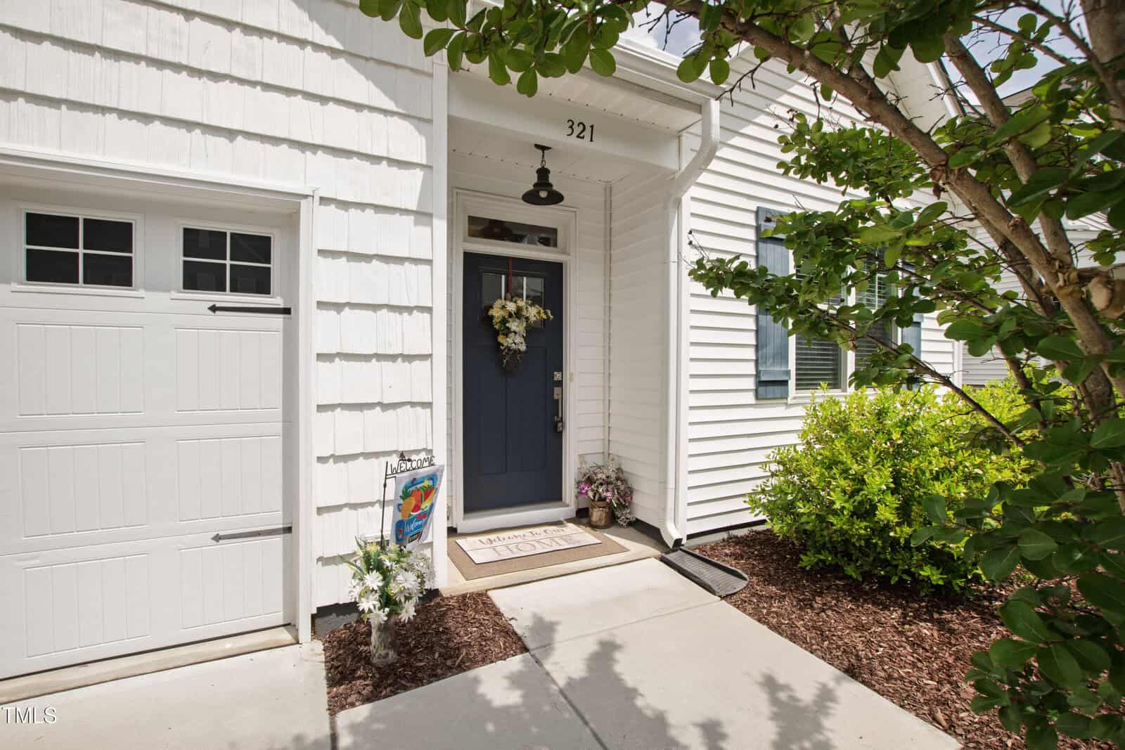 A white house exterior with a blue front door, wreath, and potted plants. The door has the number 321 above it, and a garage door is visible to the left. A tree and shrubs are on the right side.