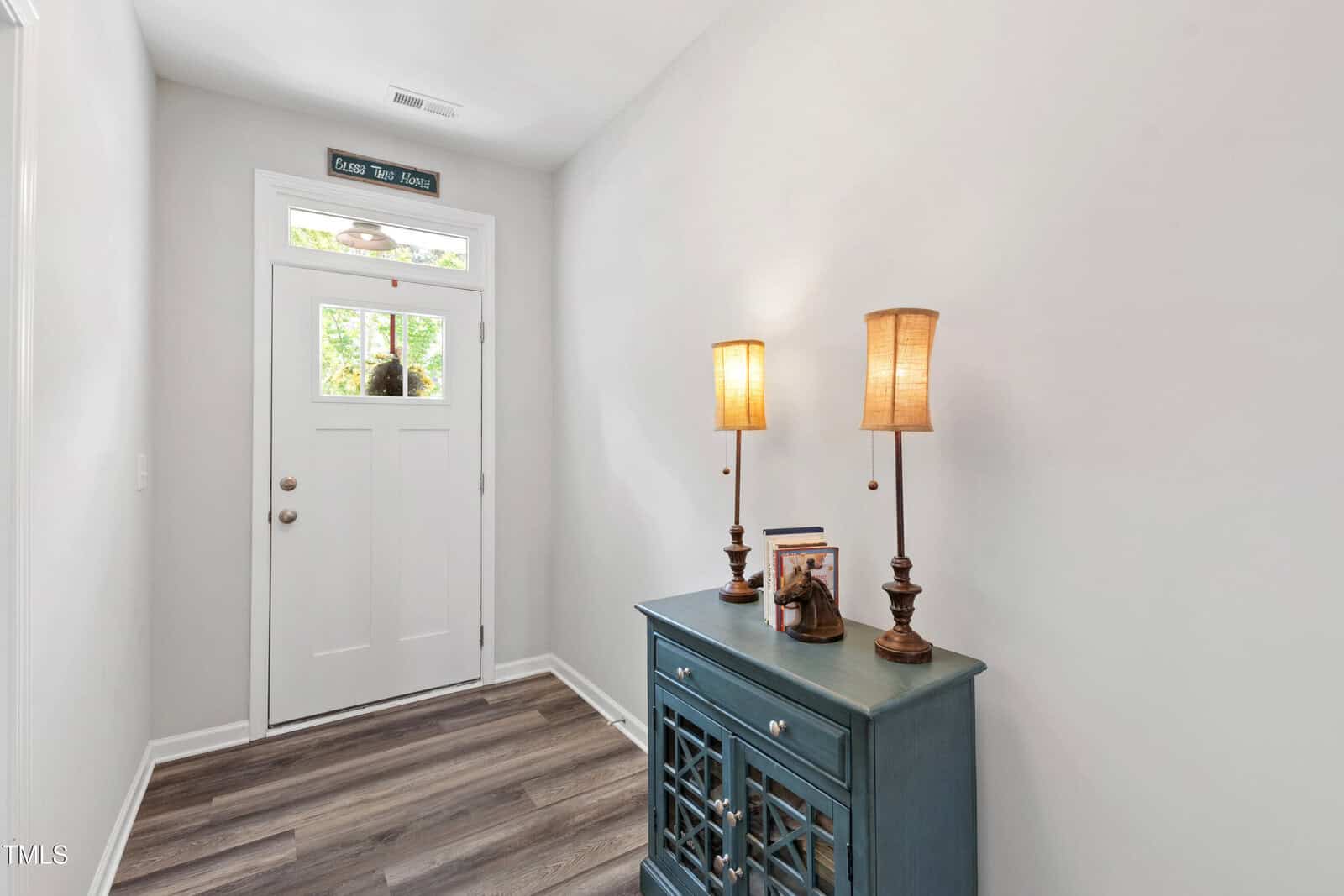 A hallway with light gray walls and wood-look flooring features a white front door with glass panels, a teal sideboard, two tall lamps, and a small decorative sign above the door that says "Come Take a Hike.