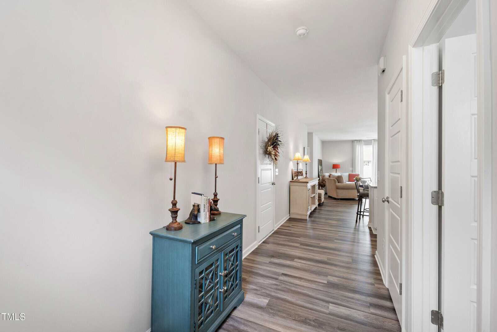 A hallway with wood-look flooring, white walls, and several white doors. A teal console table with two lamps stands on the left, leading to an open living area with couches in the background.