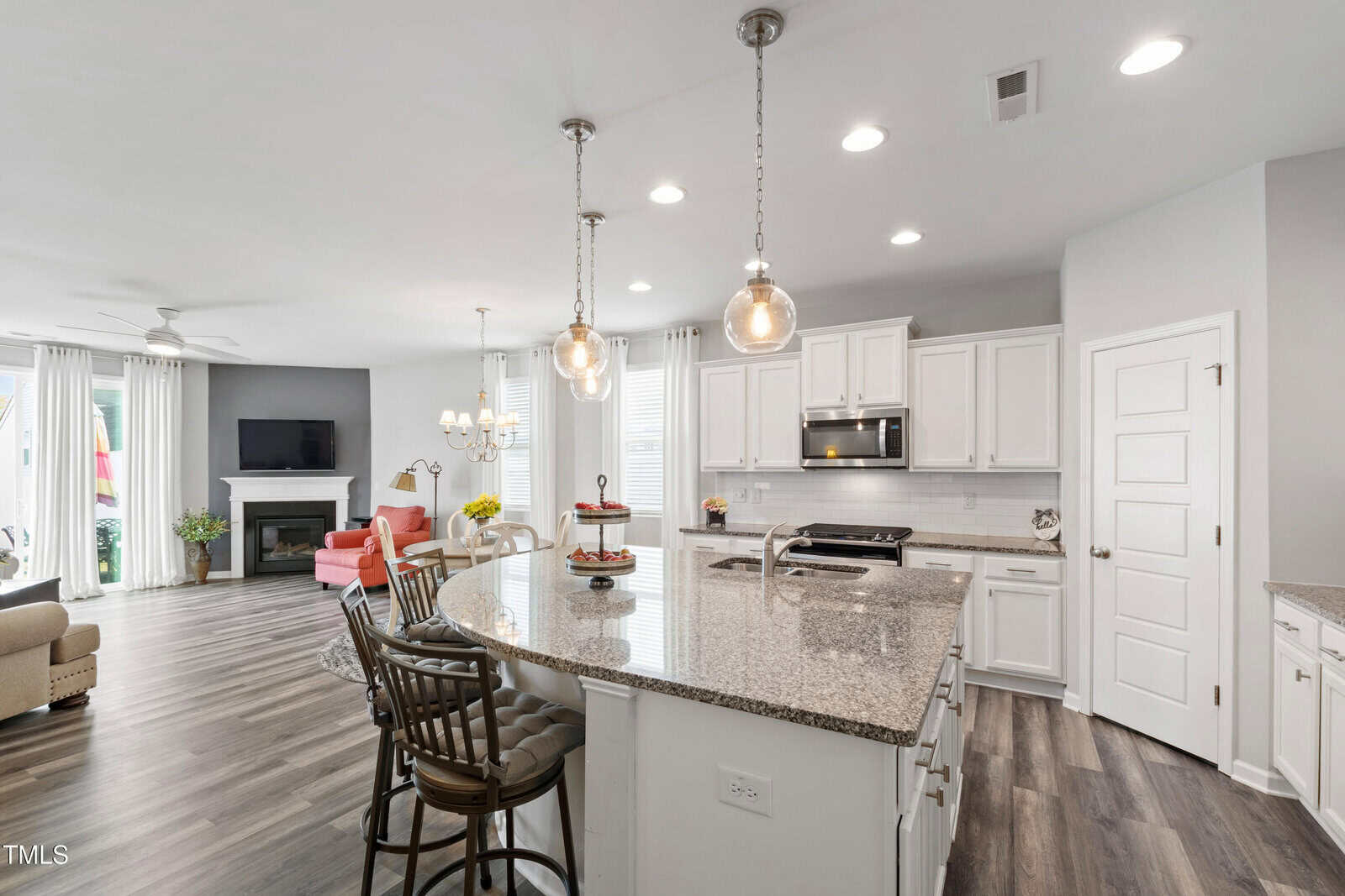 Modern kitchen with white cabinets, stainless steel appliances, granite island with bar stools, pendant lights, and an open living area with a fireplace, TV, armchairs, and large windows with white curtains.
