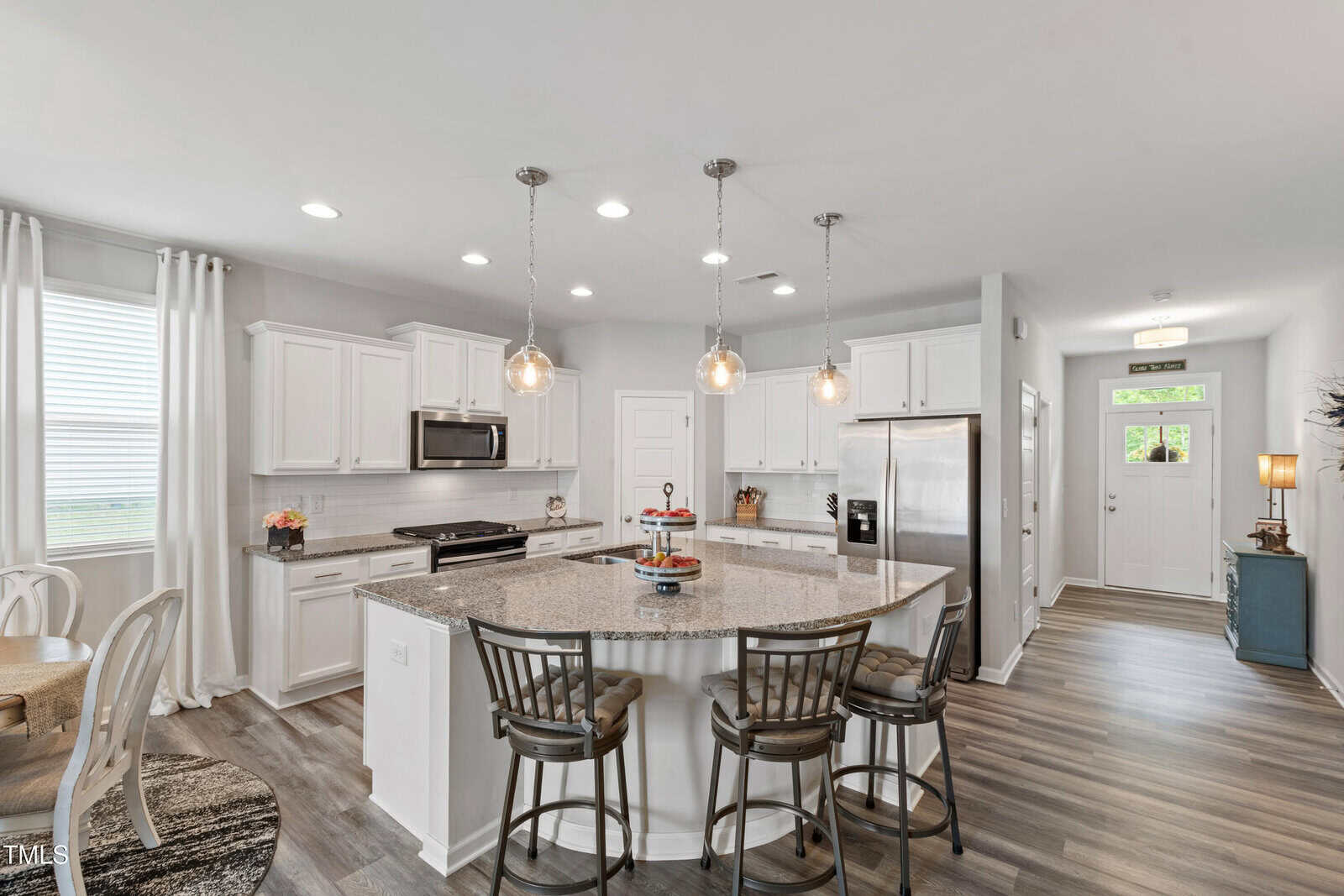 Modern kitchen with white cabinets, stainless steel appliances, granite island with three stools, pendant lights above, and wood-look flooring. Dining area with round table on the left; front door and side table visible in the background.
