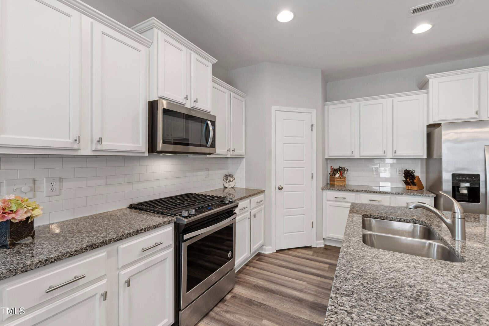 Modern kitchen with white cabinets, stainless steel appliances, granite countertops, double sink, and wood-look flooring. Subway tile backsplash and recessed lighting are visible. A small flower arrangement sits on the counter.
