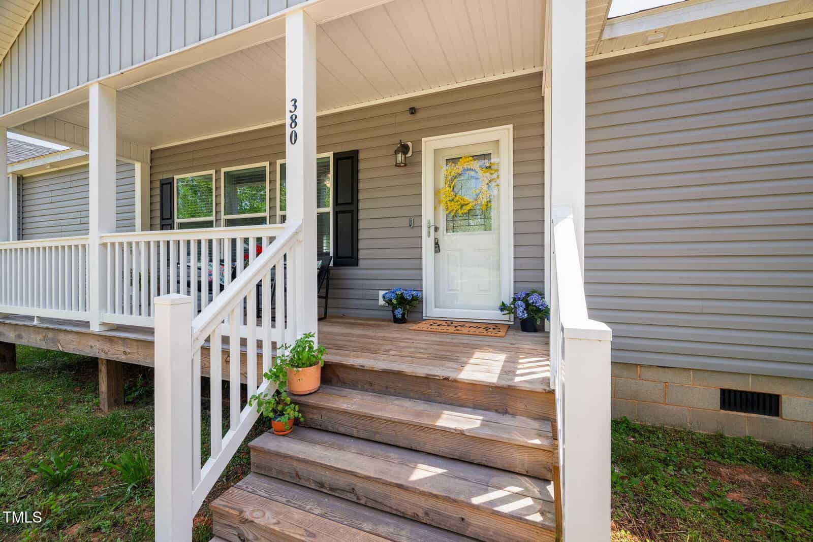 A wooden front porch with steps leading to a white door, gray siding, white railings, house number 380, potted plants on the steps, and a small welcome mat in front of the door.