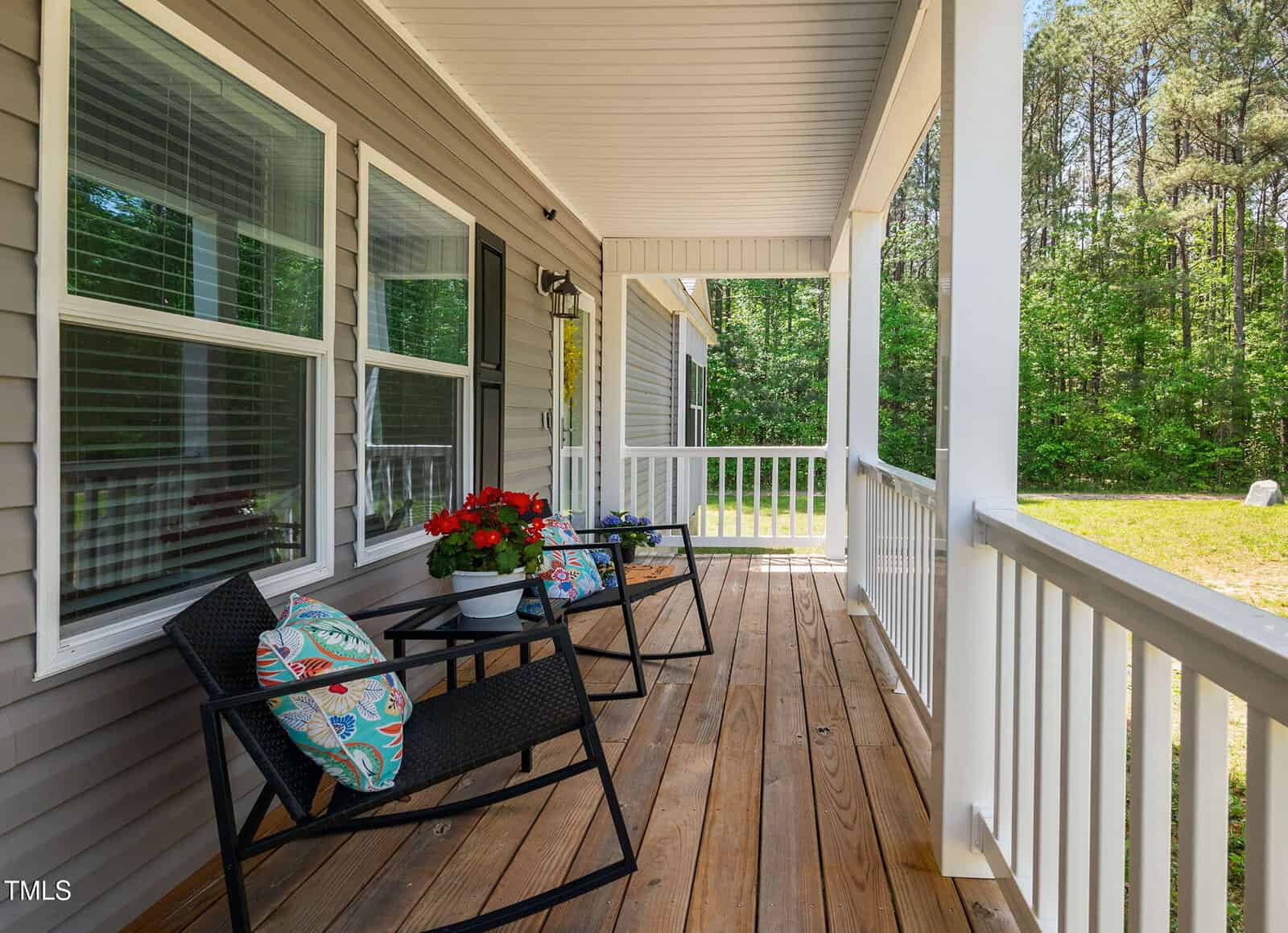 A wooden front porch with two black chairs, colorful pillows, a small table with a red flower pot, white railings, and a view of green trees and grass in the background.