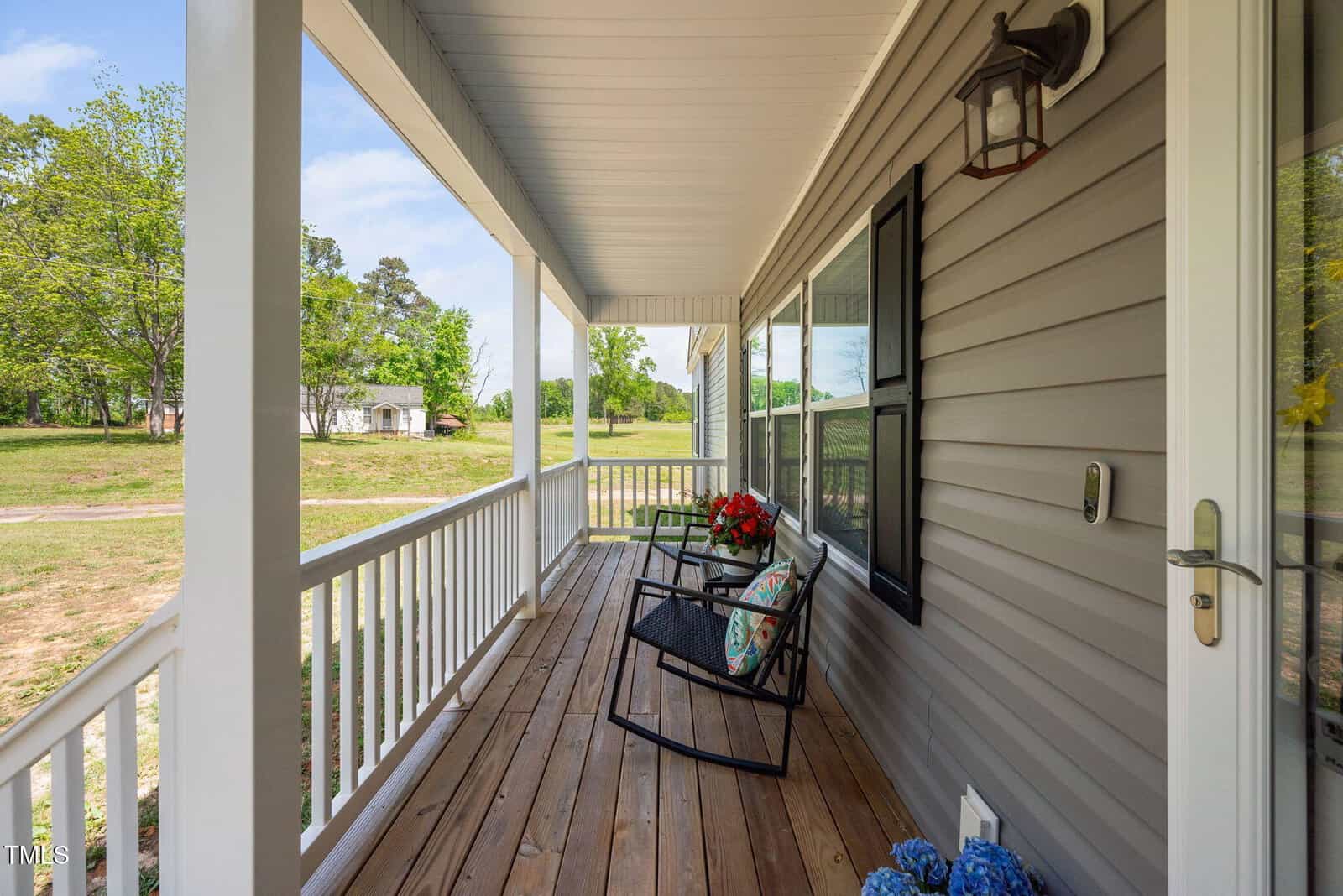 A cozy porch with a wooden floor and white railing, featuring two black rocking chairs with colorful cushions and a small table with red flowers. The porch overlooks a grassy yard with trees and a distant shed.