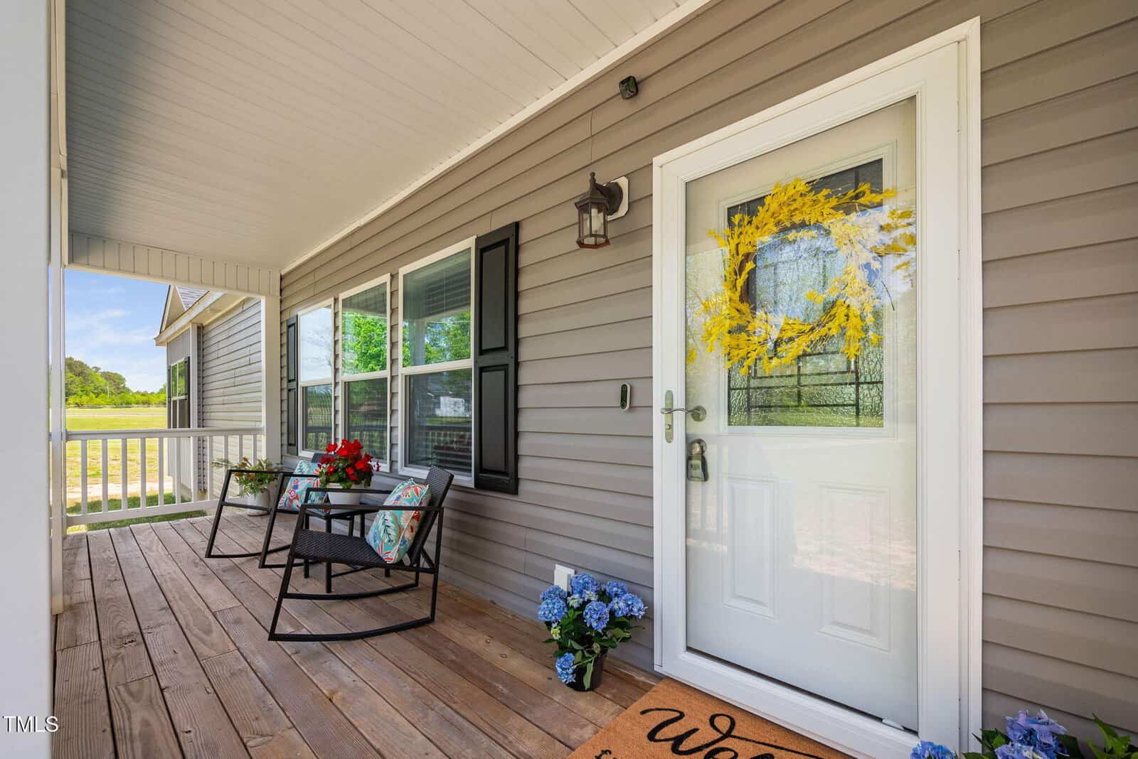 A cozy front porch with wooden flooring, two black rocking chairs with cushions and flowers, a white door with a yellow wreath, blue potted hydrangeas, and a welcome mat near the entrance.