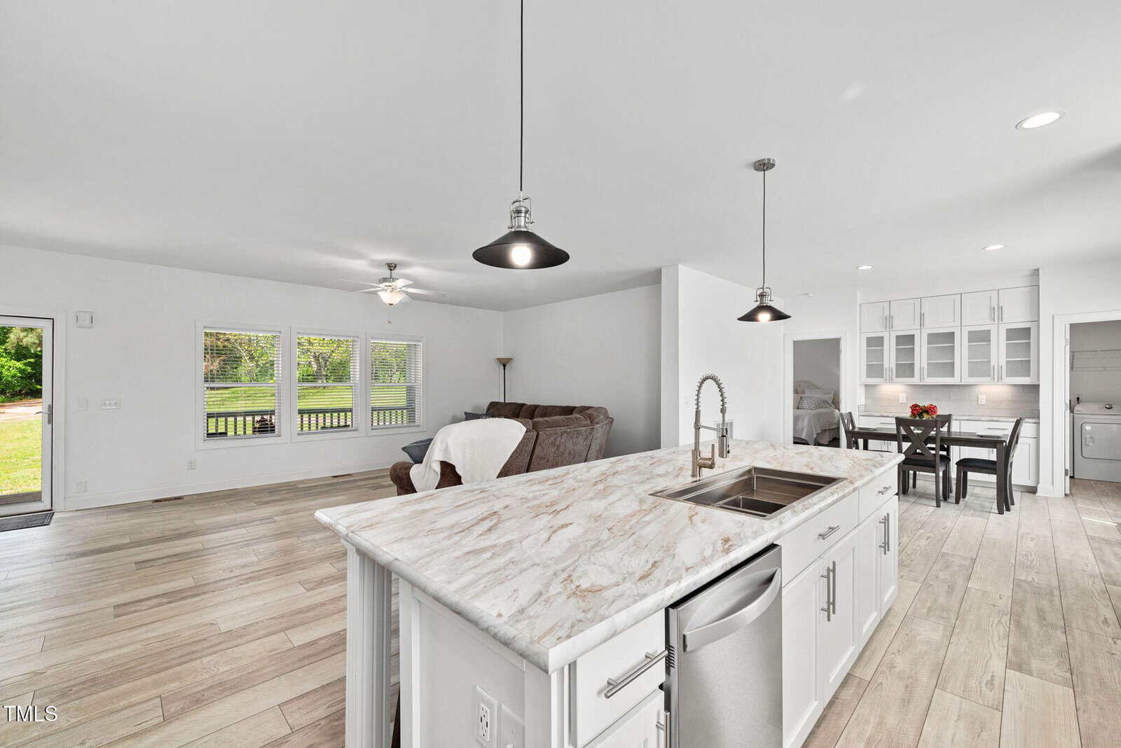 Modern open-concept living space with a kitchen island in the foreground, stainless steel appliances, light wood flooring, a sofa, dining area, and large windows providing natural light.
