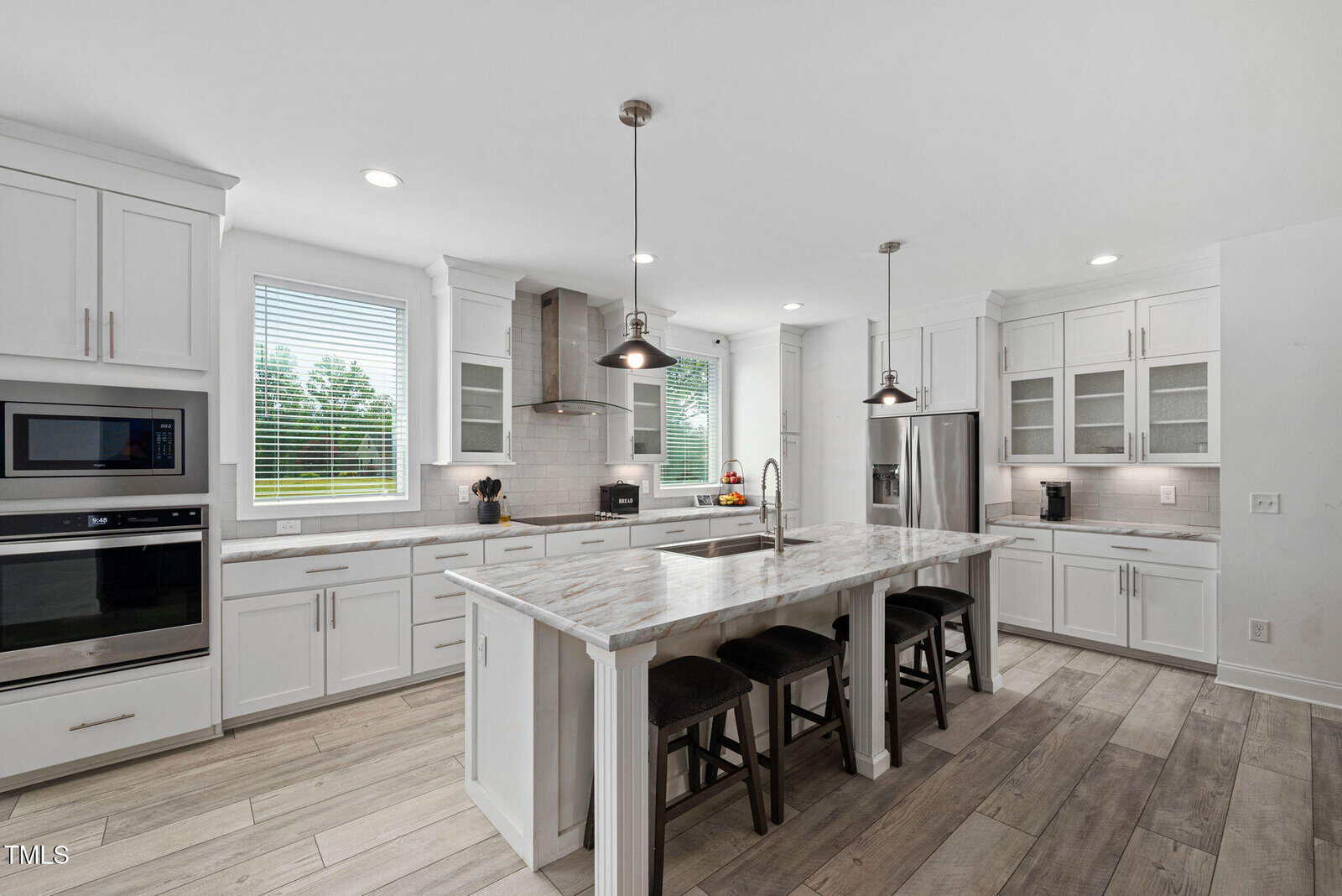 Modern kitchen with white cabinets, stainless steel appliances, marble island with three black stools, wood-look tile flooring, pendant lights, and large windows letting in natural light.