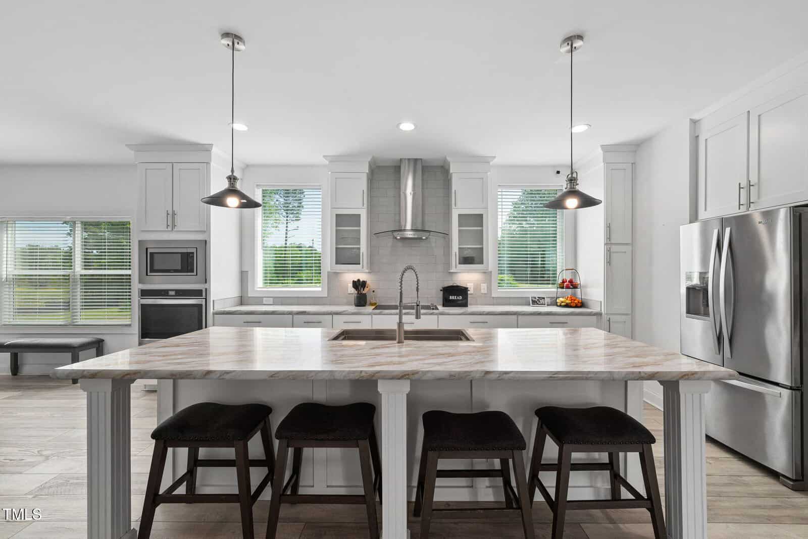 Modern kitchen with marble island, four dark stools, stainless steel appliances, two pendant lights, white cabinets, and large windows letting in natural light.