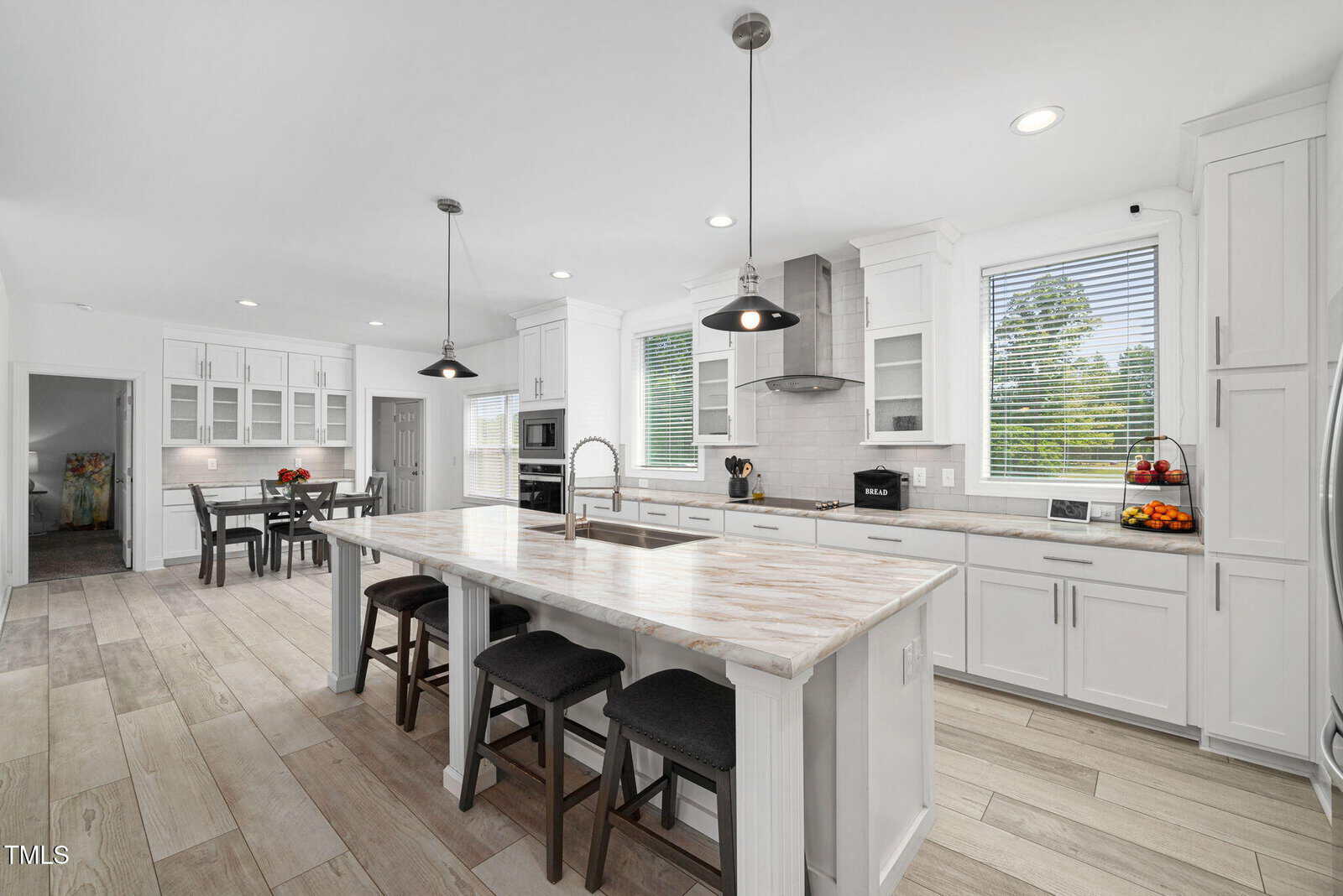 Bright modern kitchen with white cabinets, marble island with sink and stools, stainless steel appliances, pendant lights, wooden floor, and windows letting in natural light. Dining area is visible in the background.