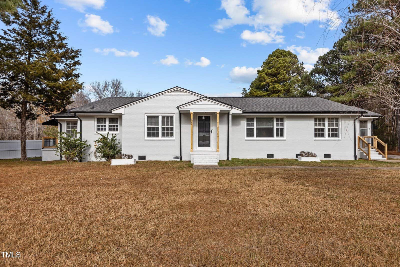 A single-story white house with black shutters at 900 Roxboro Road, Oxford, features a dark gray roof and small front steps, set on a large brown lawn surrounded by trees under a blue sky with scattered clouds.