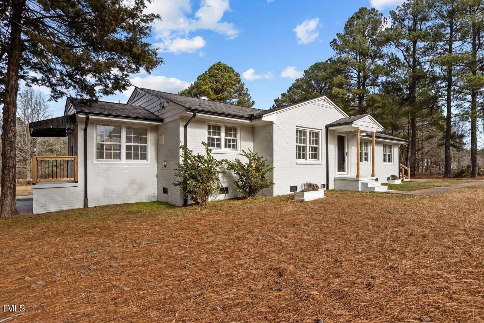 Single-story white brick house with a dark roof, large windows, and two front entrances, surrounded by pine trees and a yard covered with pine needles under a partly cloudy sky.