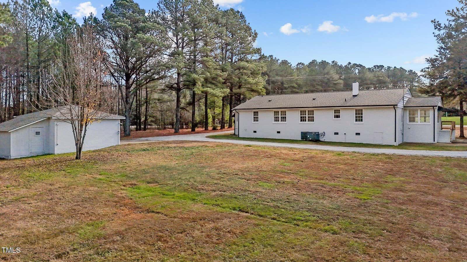 Single-story white house with a gray roof, adjacent to a detached garage. The property has a large grassy yard, surrounded by tall trees, with a paved driveway curving beside the house. Sky is clear with a few clouds.
