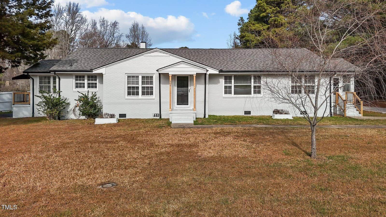 Single-story white house with a gray roof, central front door with steps, small porch, and ramps on each side. The house sits on a spacious lawn with a few small bushes and trees. Sky is partly cloudy.