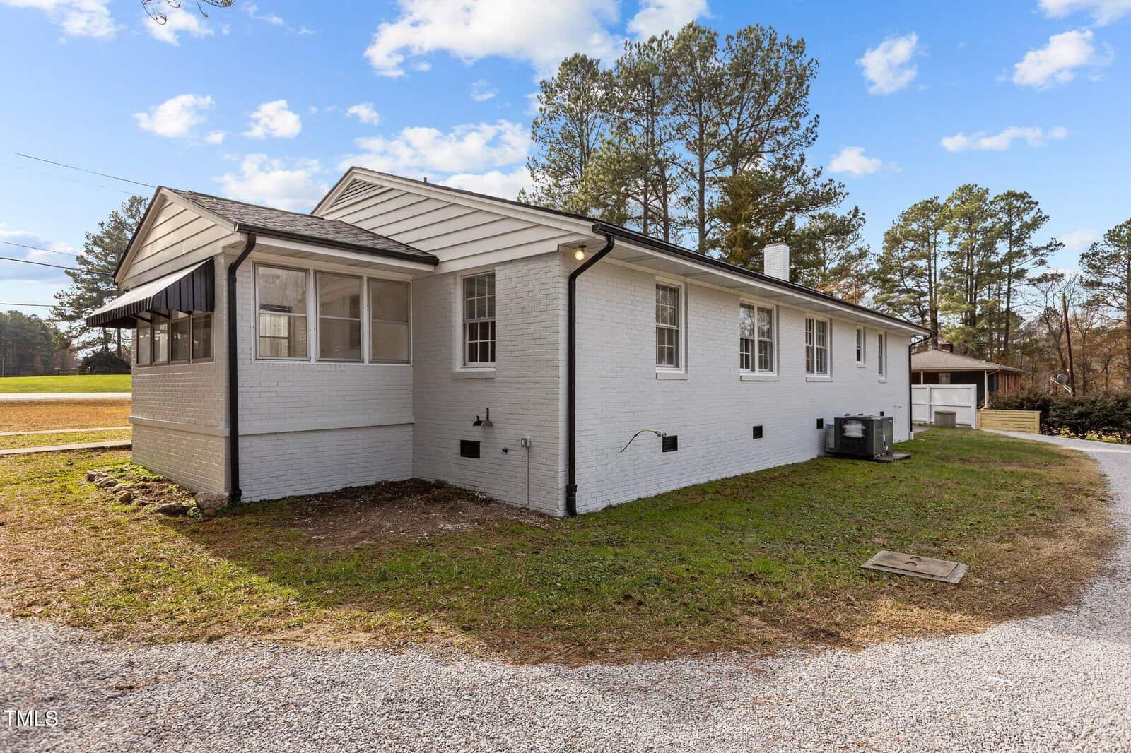 A single-story white brick house with a black roof is shown from the side. The building has multiple windows, a small enclosed porch, and is surrounded by grass, gravel, and trees in the background.