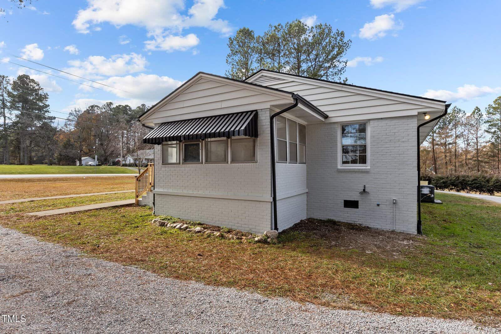 A small, single-story white brick house with a black awning over the front window, white siding, and a gravel driveway. The yard has patchy grass and is surrounded by trees under a partly cloudy sky.