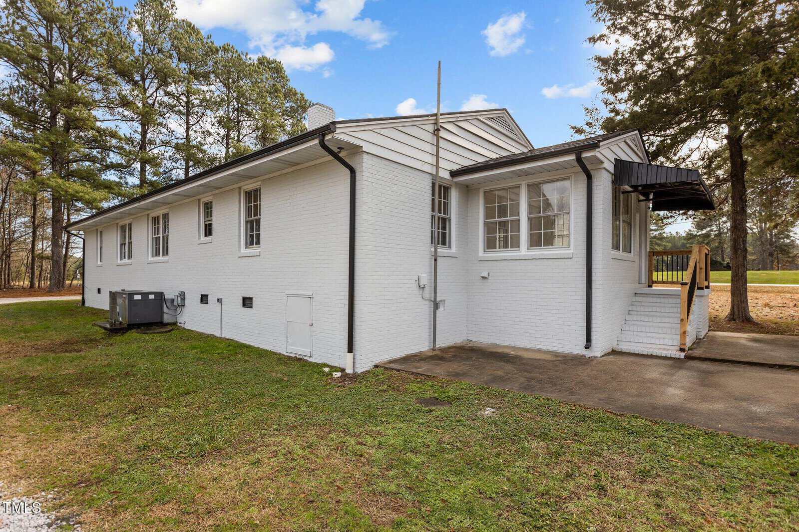 A single-story white brick house with multiple windows, a small set of stairs with a black awning over the entrance, an outdoor air conditioning unit, and a grassy yard surrounded by trees.