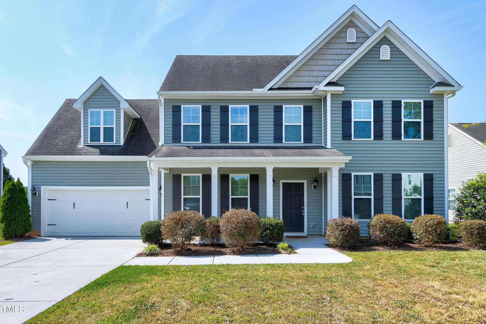 Two-story blue house with white trim, black shutters, and a covered front porch. There is a double garage on the left and neatly trimmed bushes along the front. The lawn is green, with a clear blue sky in the background.