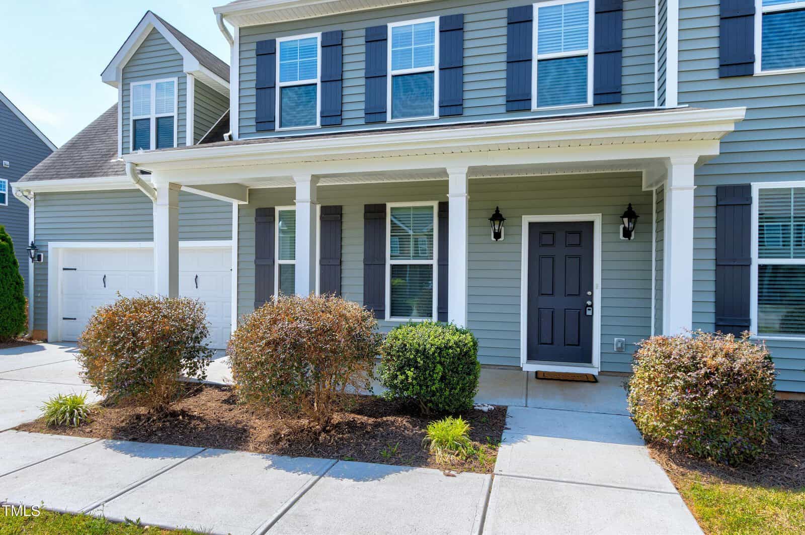 Two-story house with light blue siding, white trim, black shutters, and a covered front porch. There are two garage doors, shrubs along the walkway, and multiple windows on both floors.