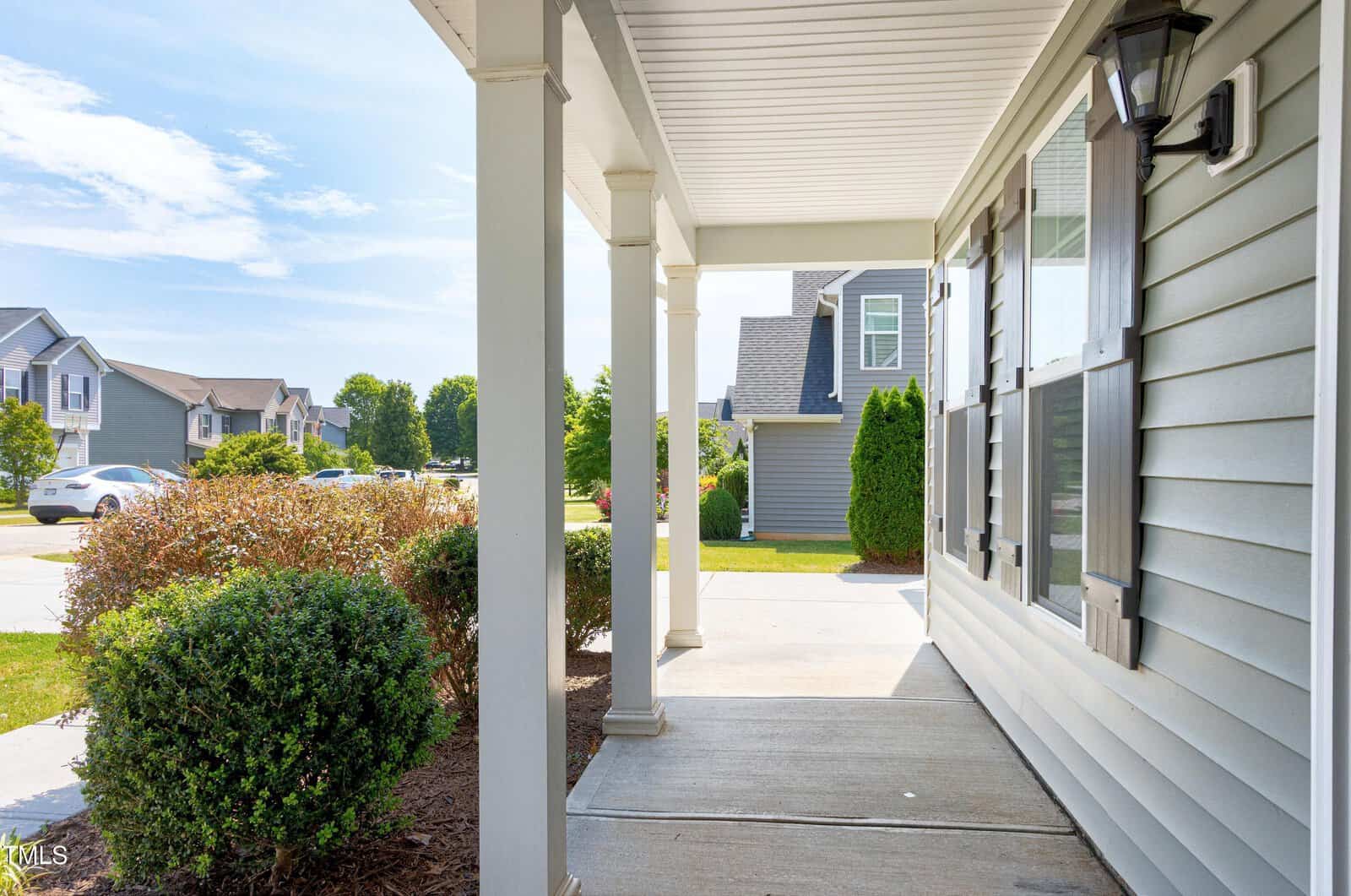 Covered front porch of a suburban home with light gray siding, black shutters, and white columns. Bushes line the walkway, and neighboring houses are visible in the background on a sunny day.