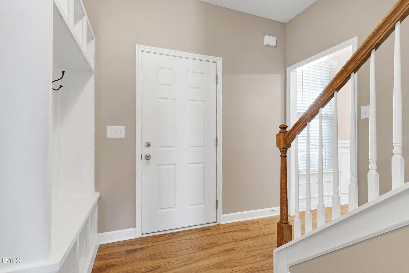 A bright entryway with a white front door, light tan walls, wood flooring, a wooden staircase with white railings, and a built-in bench with coat hooks on the left. Natural light streams in from an adjacent room.