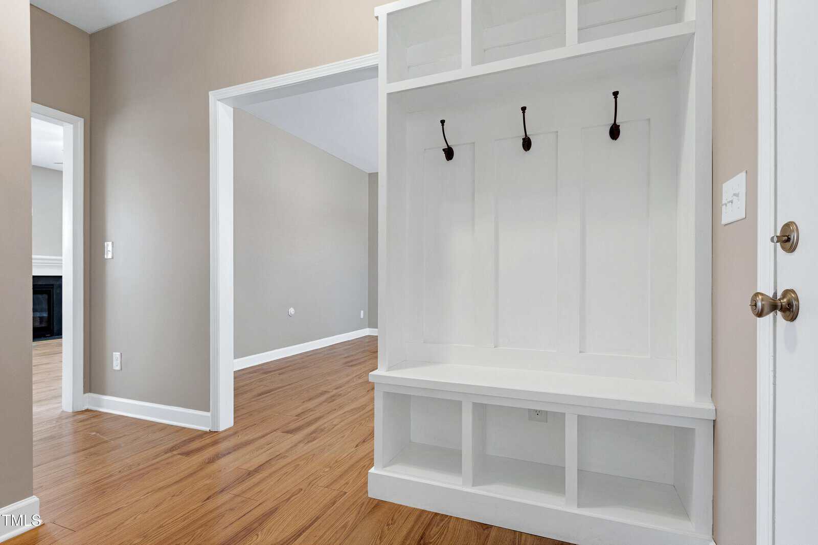 A white built-in entryway bench with storage cubbies and three black wall hooks is set against beige walls in a hallway with wood flooring. Open doorways lead to adjoining rooms.