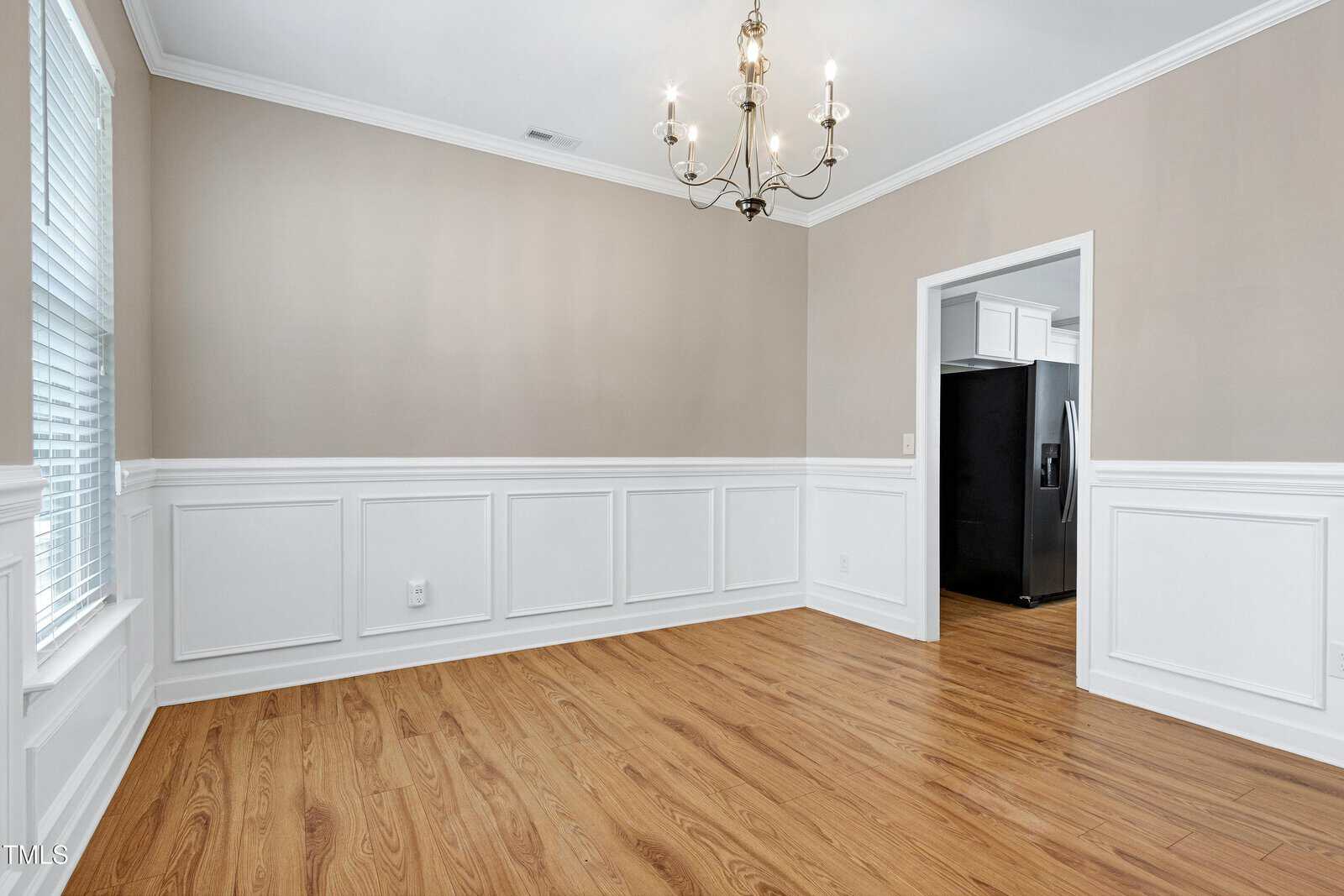 Empty dining room with light brown wood flooring, white wainscoting, beige upper walls, a chandelier, large window with blinds, and an open doorway leading to a kitchen with visible refrigerator.