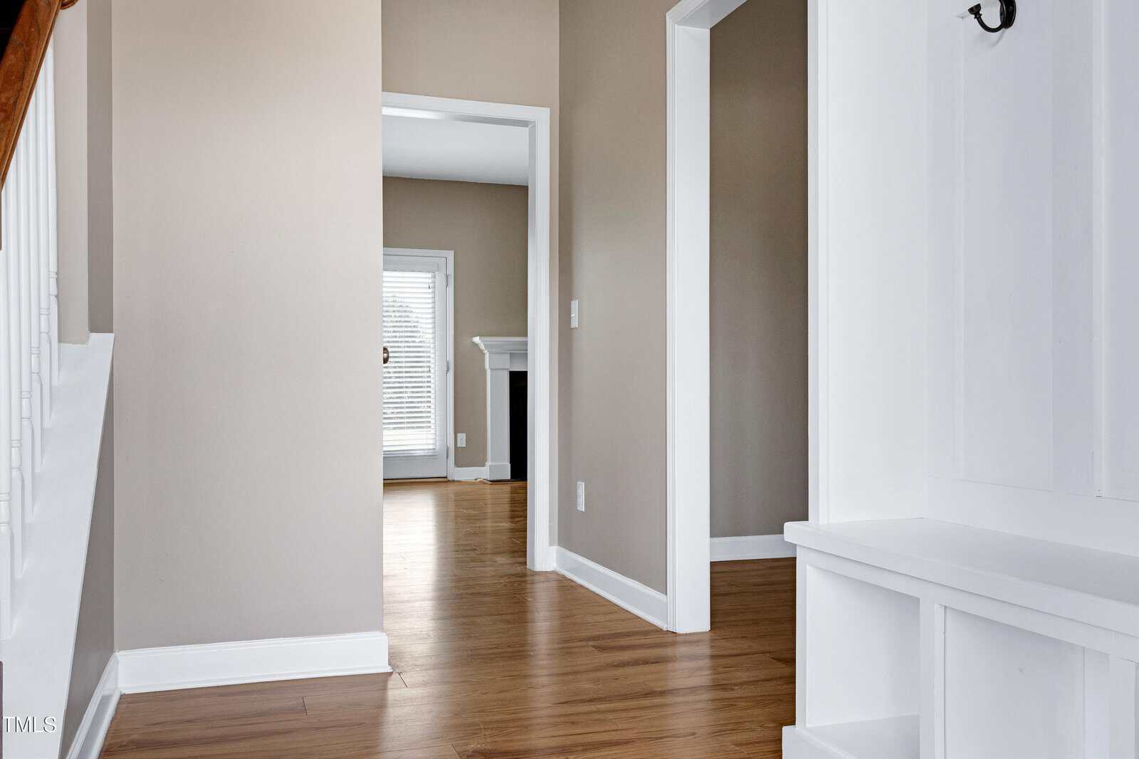 A hallway with light brown walls, white trim, and wood flooring leads to another room with a fireplace and tall window blinds. A built-in white bench with hooks is on the right.