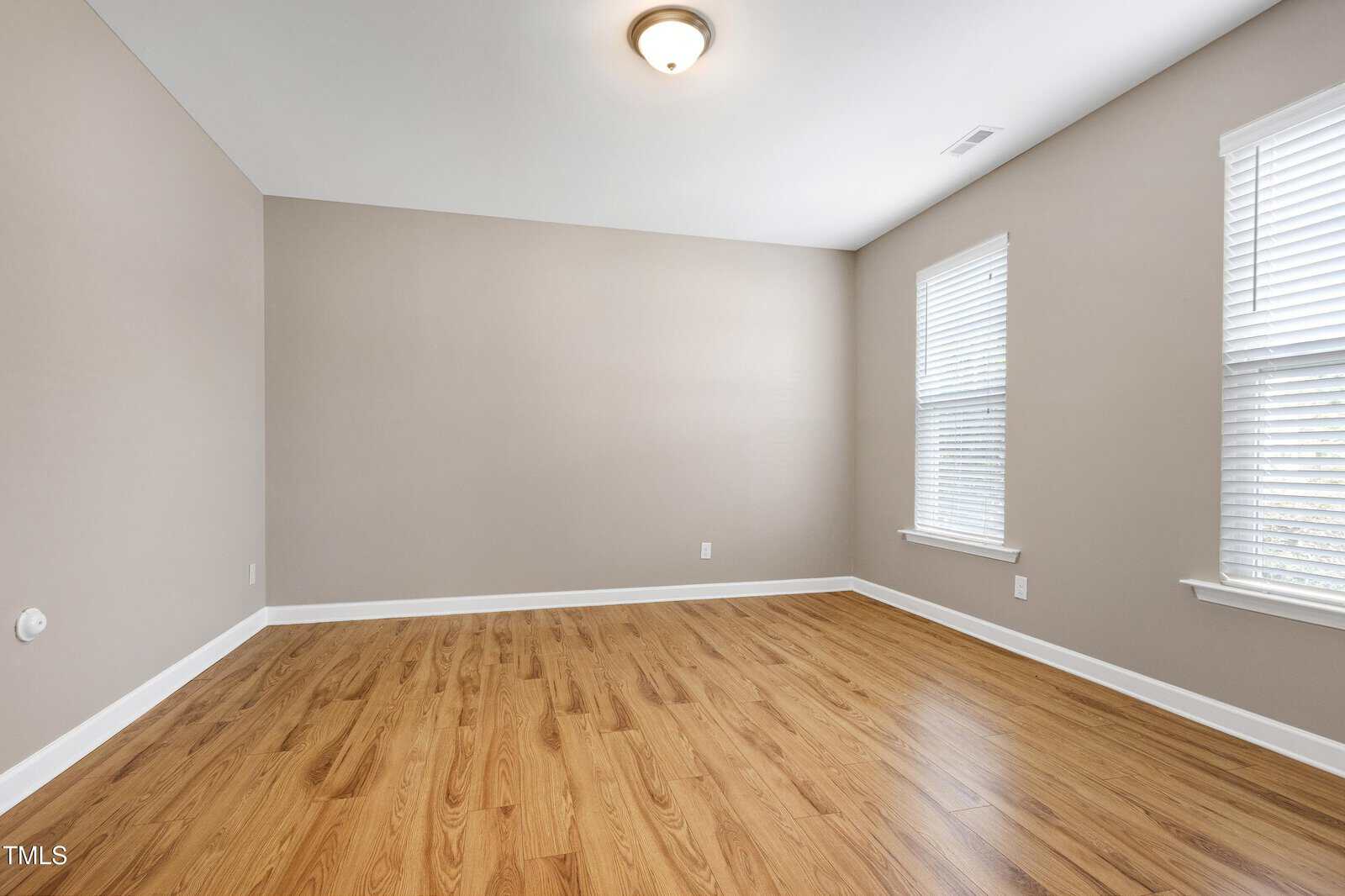 Empty room with beige walls, white trim, two large windows with blinds, hardwood flooring, and a ceiling light fixture. No furniture or decorations are present.