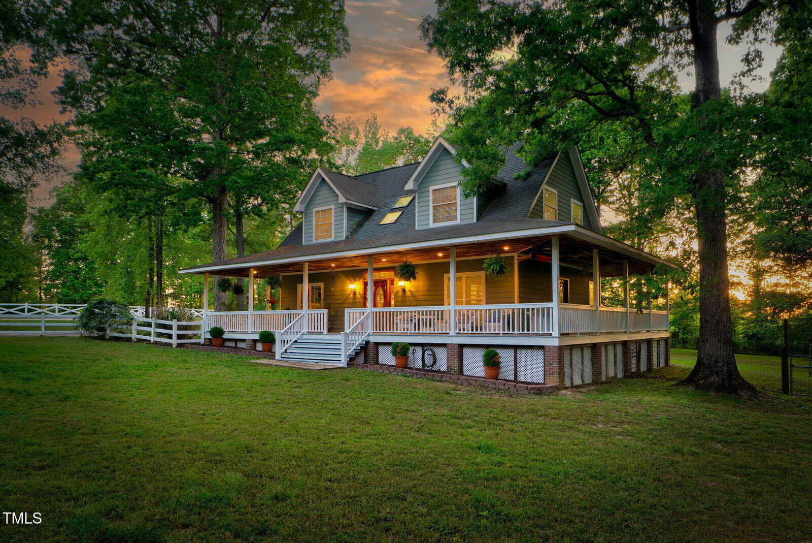 A two-story country house with a wraparound porch, white railings, and large windows sits among tall trees at sunset, with warm lights glowing inside and a grassy yard in front.