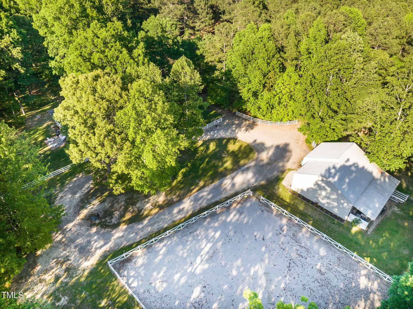 Aerial view of a rural property with a white-fenced riding arena, a barn or shed, gravel paths, and dense green trees surrounding the area. Sunlight casts long shadows across the scene.