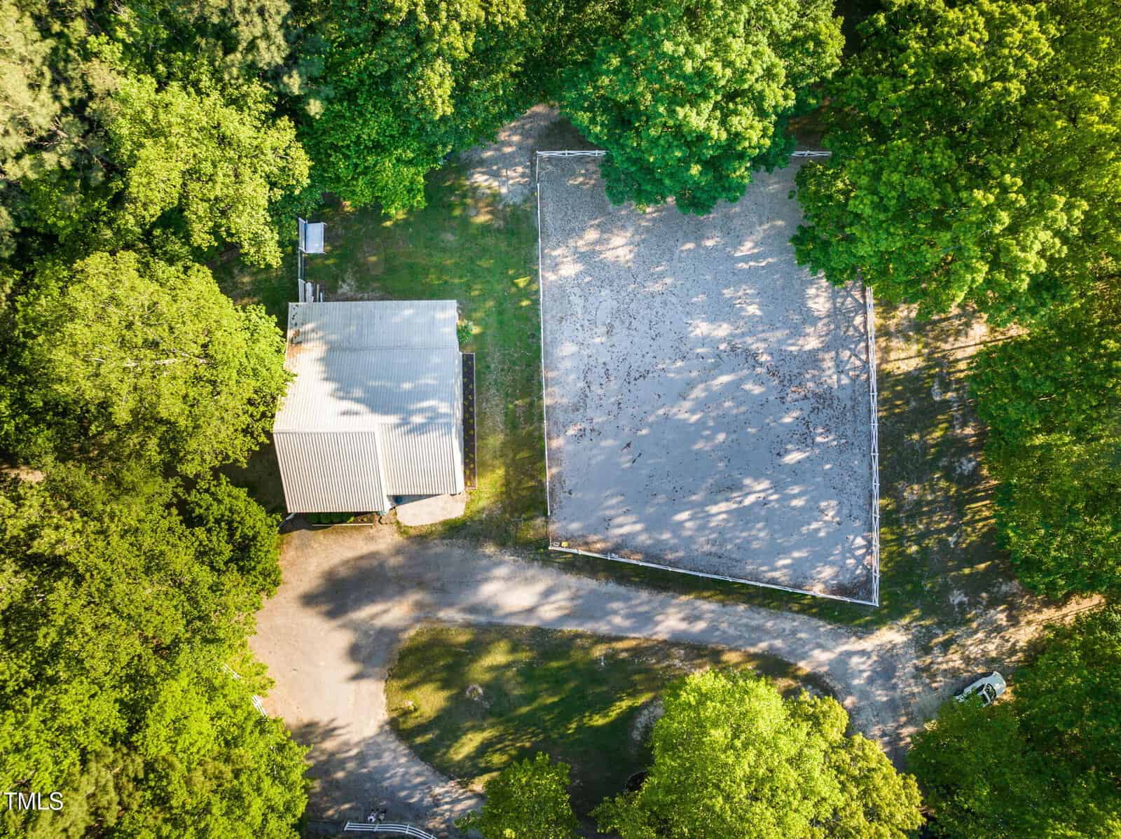 Aerial view of a small building with a gray roof next to a large rectangular sand or gravel area, surrounded by green trees and partially shaded, with a dirt path leading through the scene.