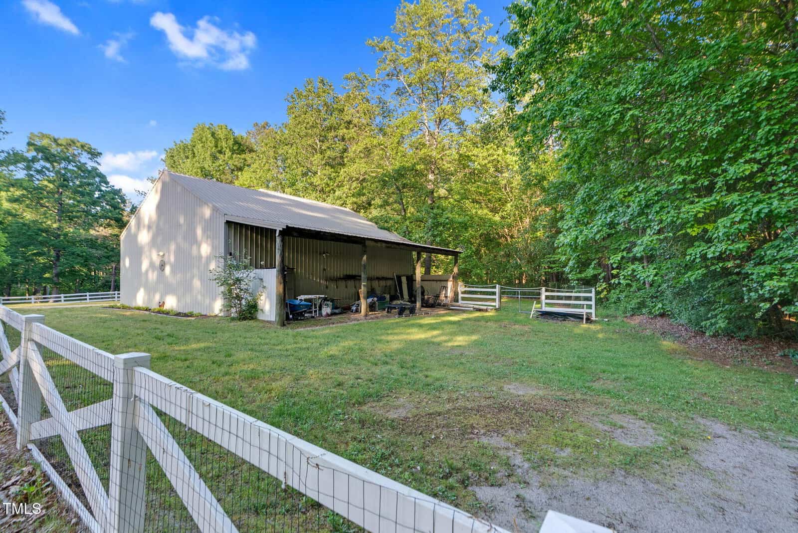 A white barn with a metal roof stands in a grassy, fenced yard surrounded by trees. The barn has an open side with tools and equipment visible inside, and sunlight shines through the trees.