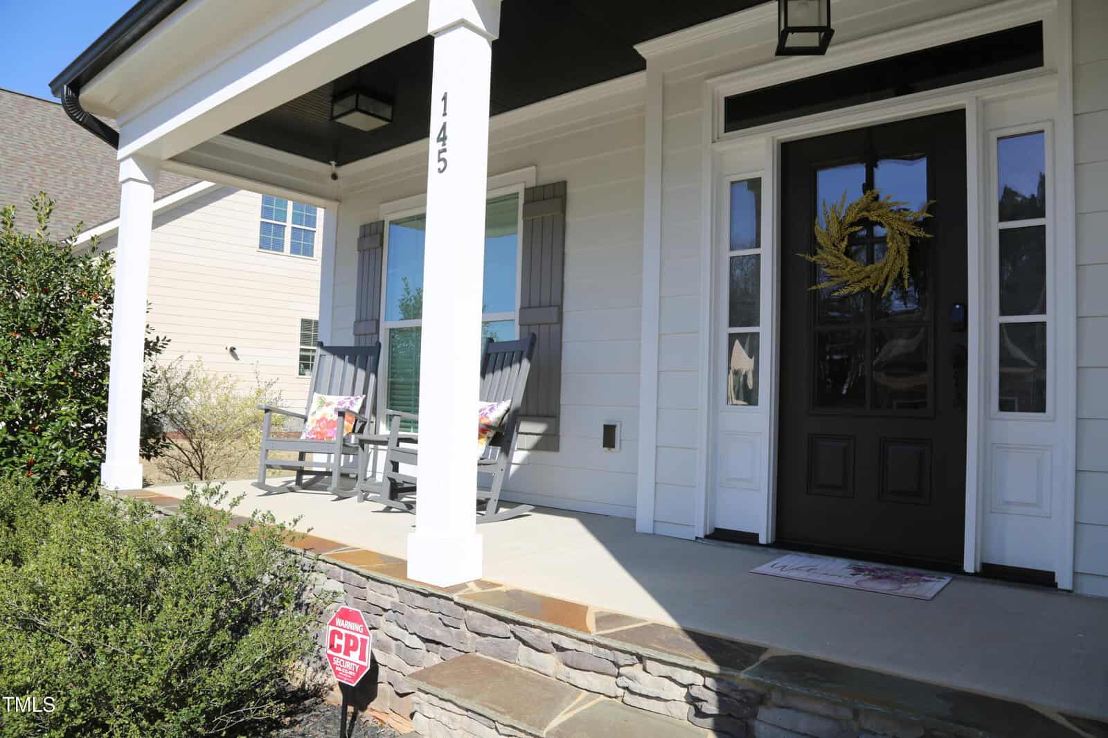 Front porch of a white house with two rocking chairs, a mat at the door, a wreath on the black front door, and a CPI security sign in the garden. The porch has columns, stone accents, and green bushes in front.