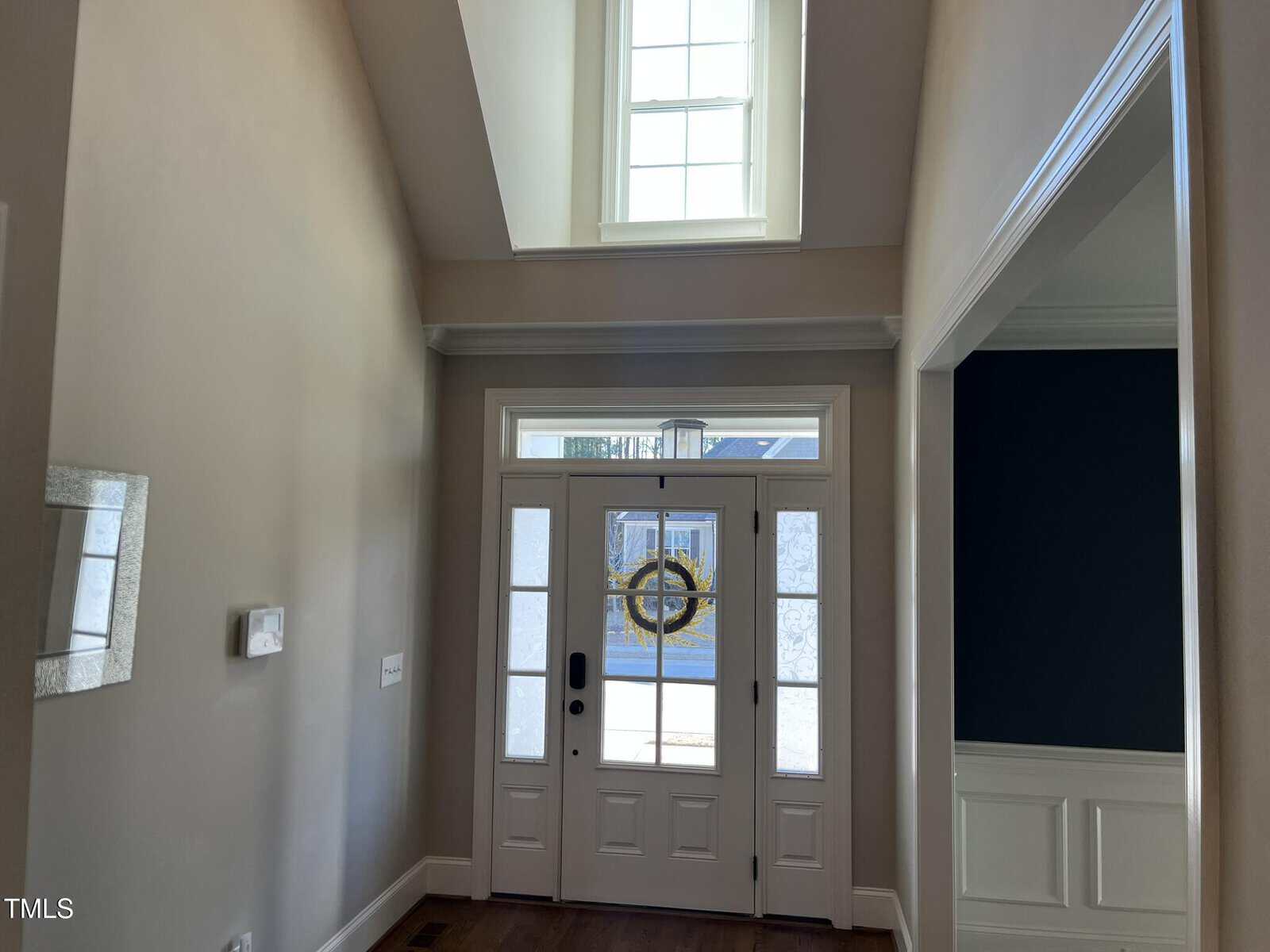 A foyer with double front doors featuring glass panels, a large transom window above, beige walls, wood flooring, and a view of a dark blue room with white trim on the right. Natural light enters from the windows.