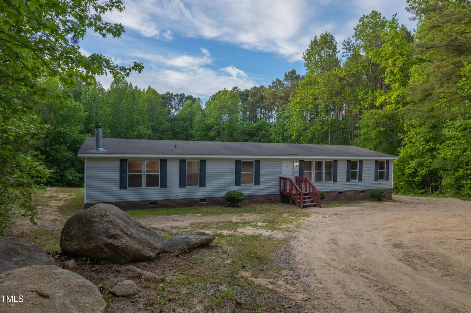 A single-story manufactured home with gray siding and black shutters sits on a large dirt lot, surrounded by trees and large rocks under a partly cloudy sky. A small red staircase leads to the front door.