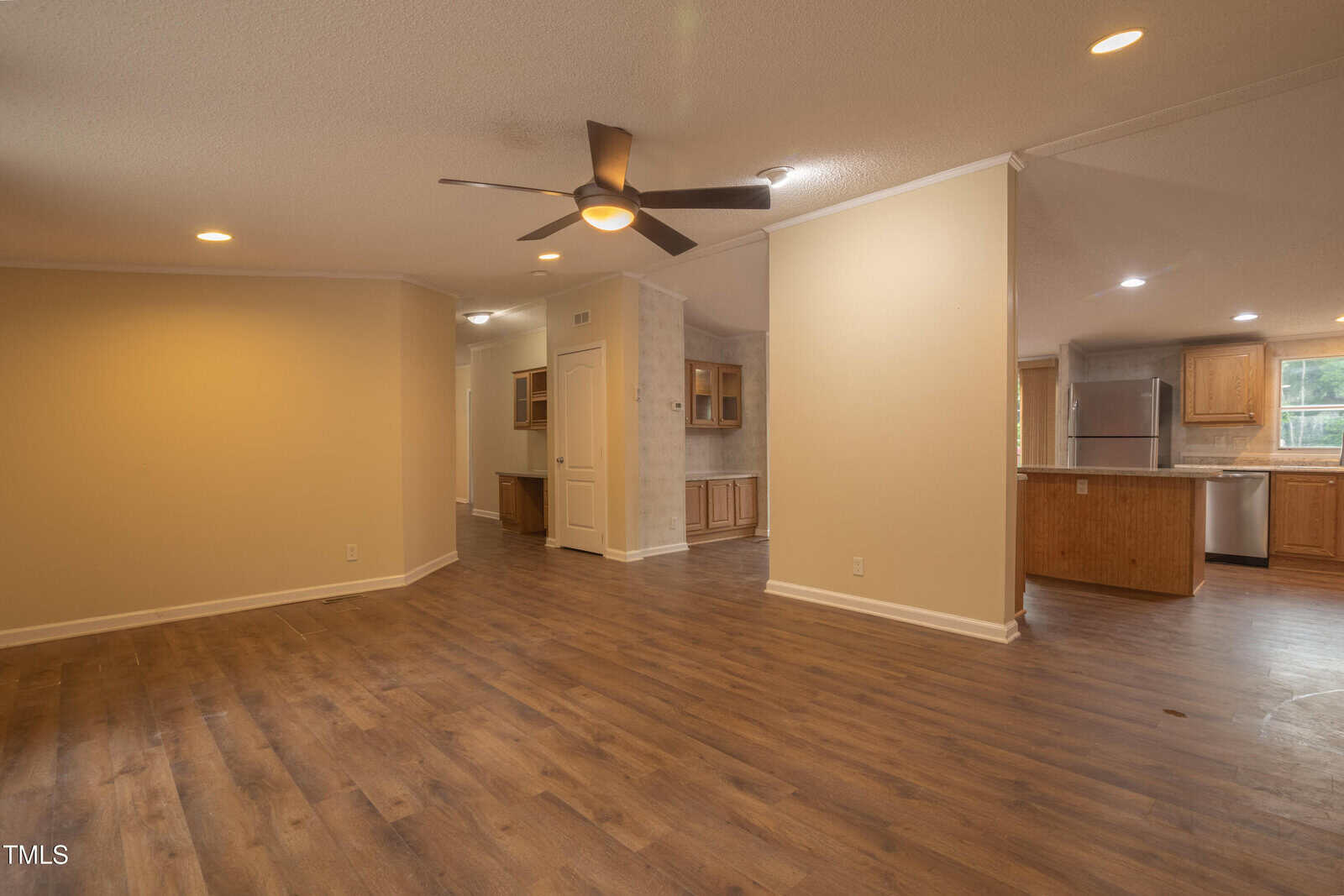 Spacious open-concept living area with wood-look flooring, beige walls, ceiling fan, and recessed lighting. Kitchen with stainless steel appliances and wood cabinets visible in the background.