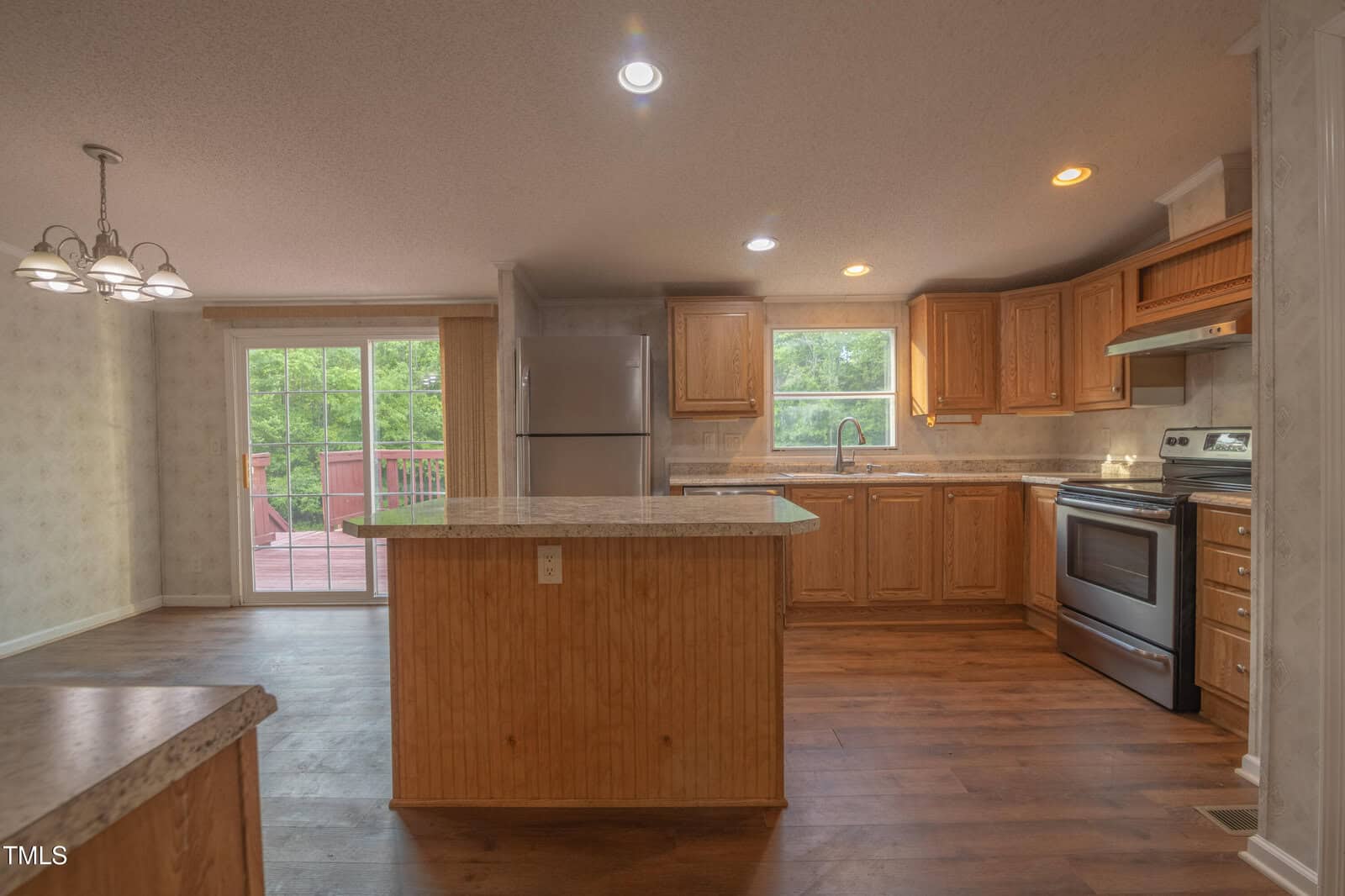 A kitchen with wooden cabinets, stainless steel appliances, a central island, and hardwood floors. Large windows and a sliding glass door provide natural light and a view of a deck and trees outside.