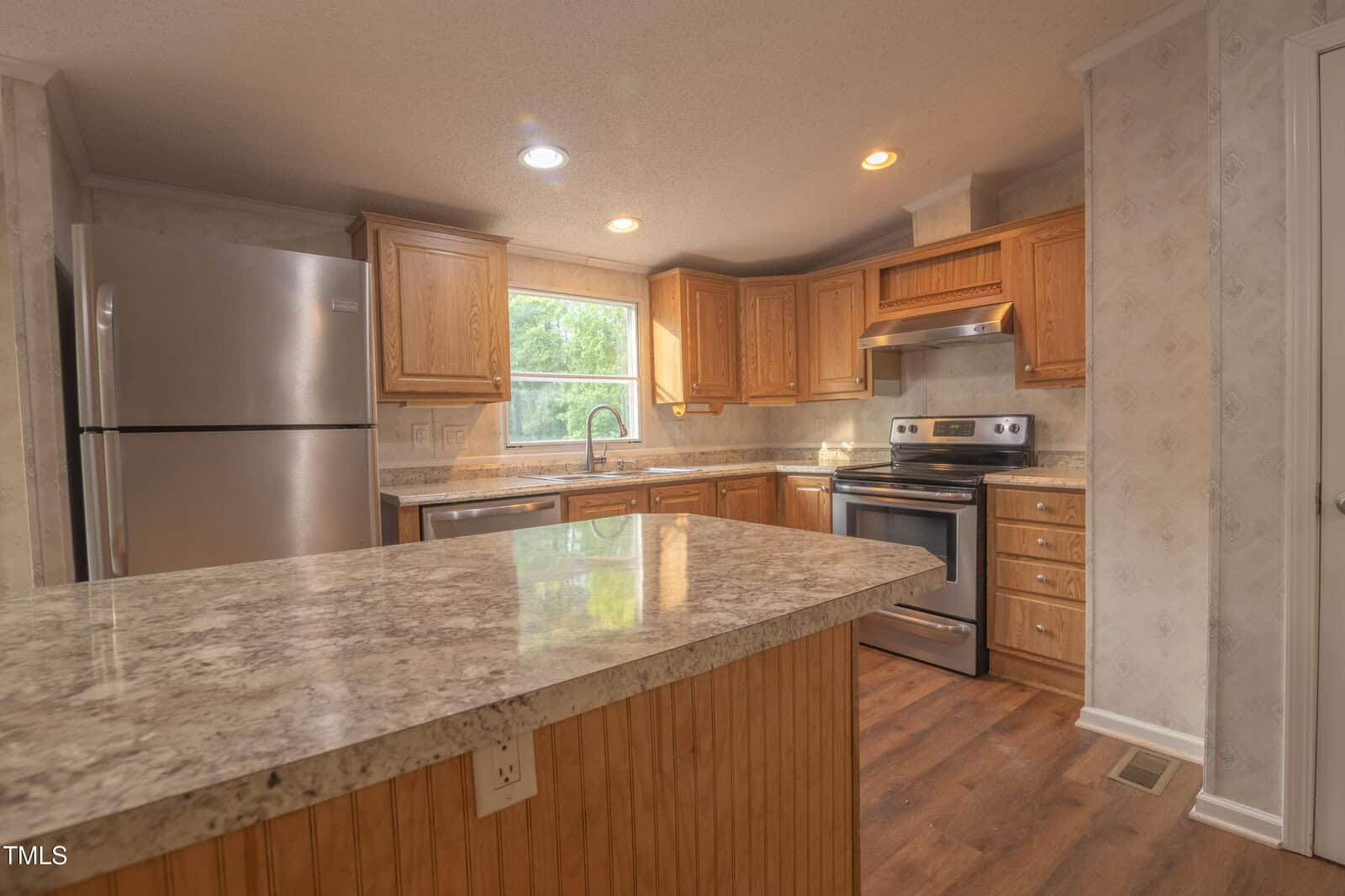 Modern kitchen with stainless steel refrigerator and stove, wooden cabinets, granite countertops, a large island in the foreground, and wood-style flooring. A window is above the sink, letting in natural light.