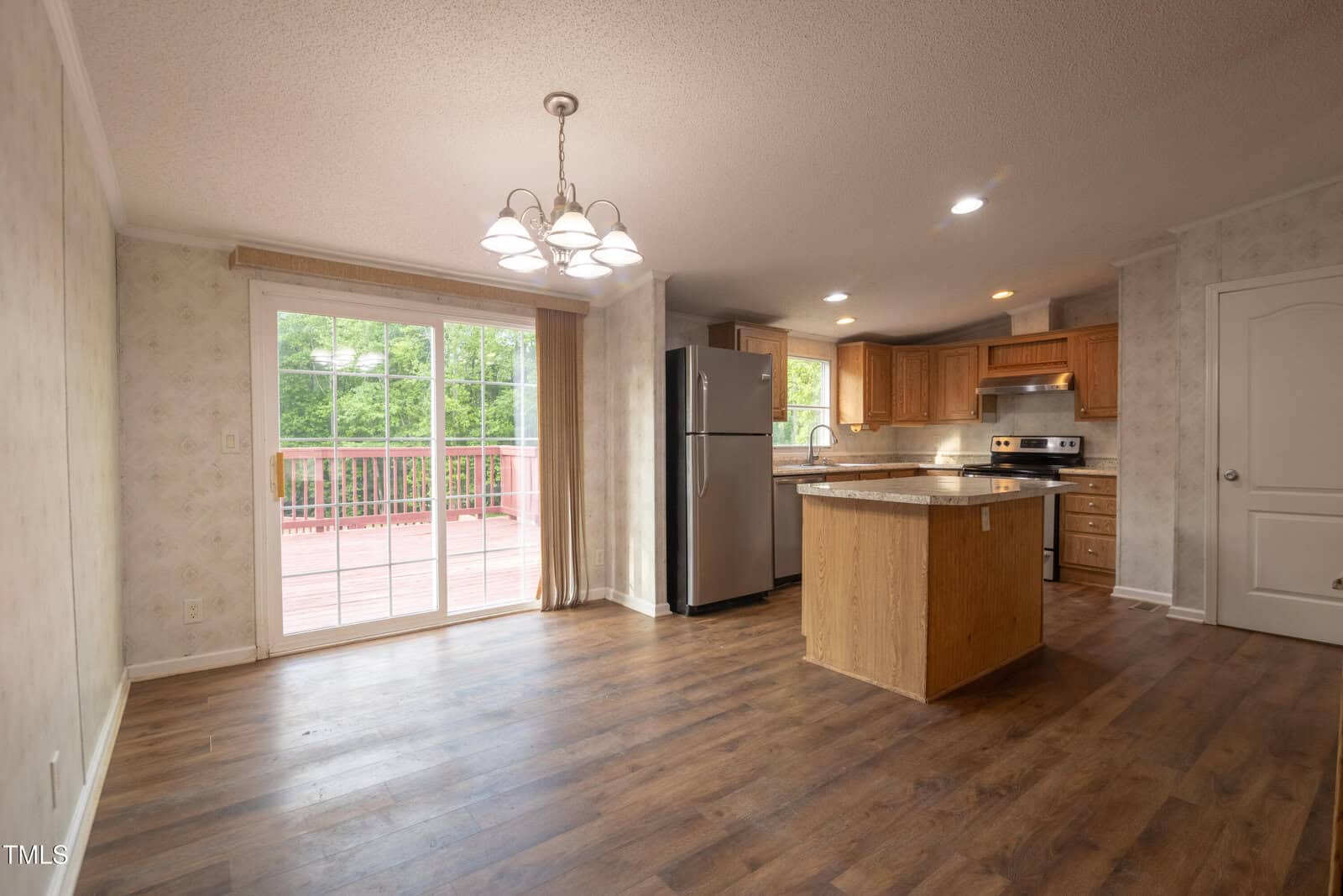 A kitchen and dining area with wood flooring, stainless steel appliances, wooden cabinets, a center island, and a chandelier. Large sliding glass doors open to a red deck with a view of trees outside.