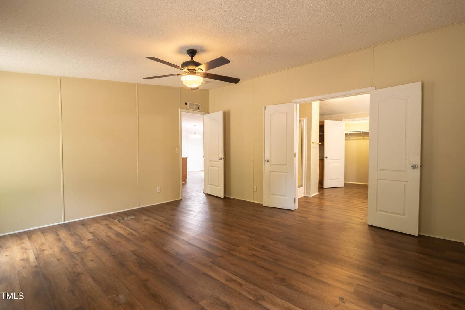 Empty room with wood flooring, beige walls, ceiling fan with light, and double doors leading to a walk-in closet. Natural light enters from an open doorway on the left side.