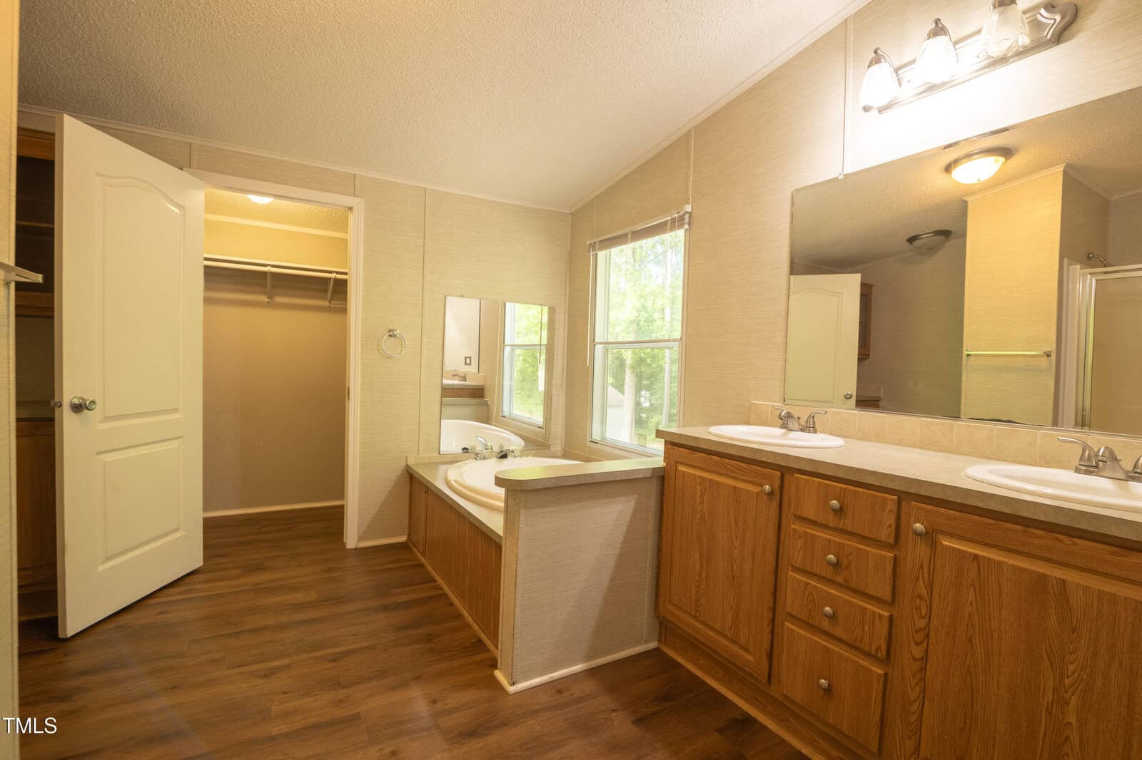 Spacious bathroom with wood flooring, double sink vanity, large mirror, bathtub next to window, and open door leading to a walk-in closet. Bright lighting and neutral wall colors create a warm atmosphere.