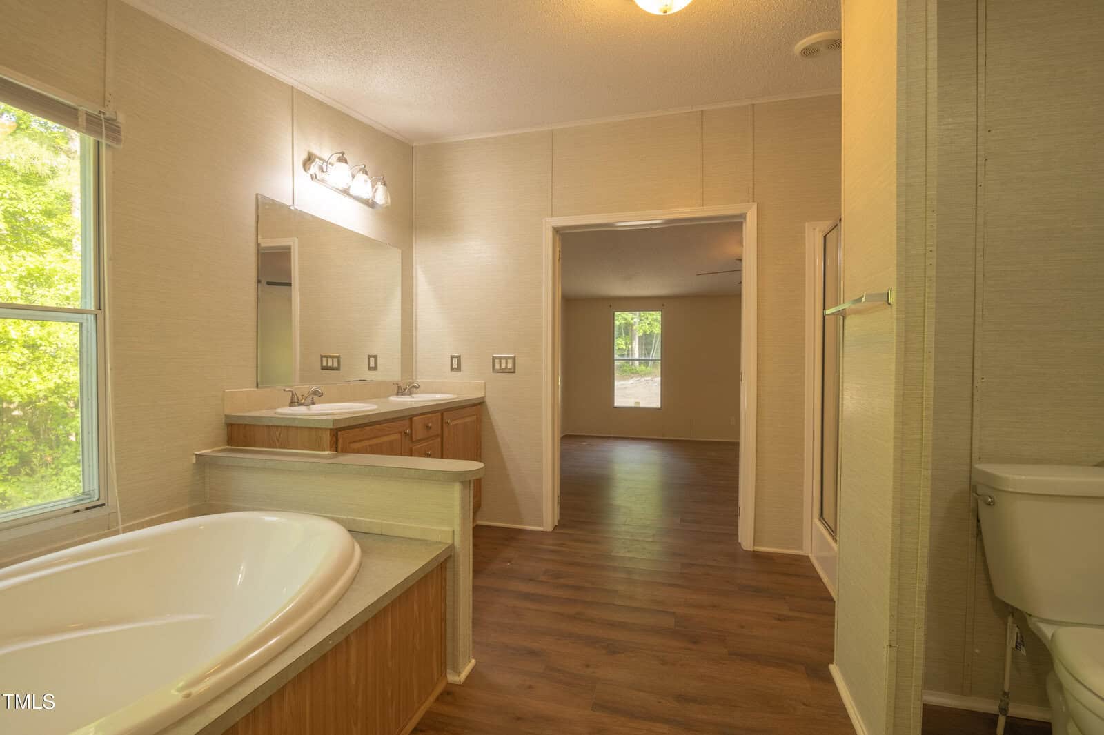 Bathroom with a large soaking tub, double sink vanity, toilet, and wood-look flooring. The room opens to a space with a window and view of greenery outside.