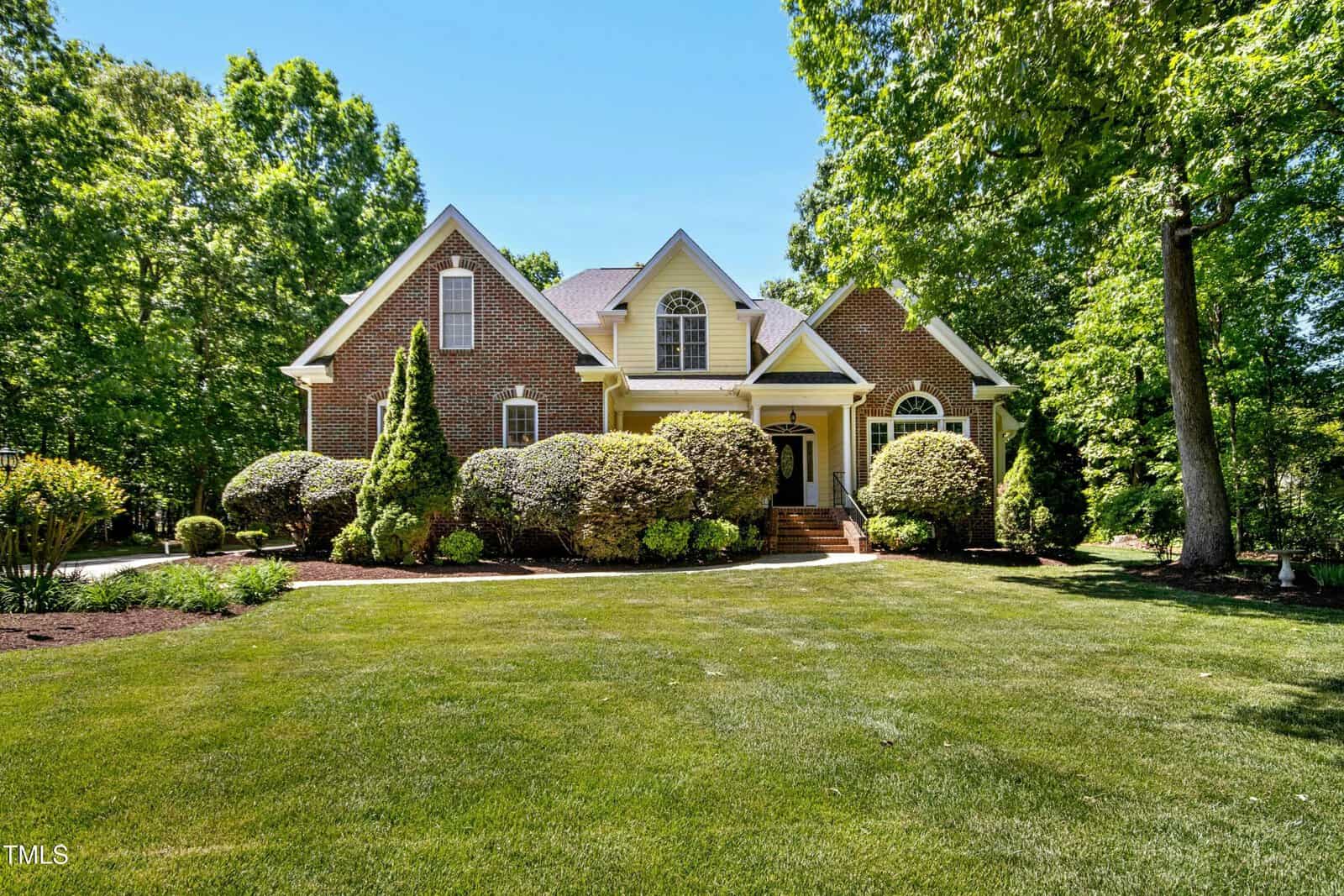 A large brick house with white trim and tall windows, surrounded by neatly trimmed bushes and mature trees, sits on a well-manicured lawn under a clear blue sky.