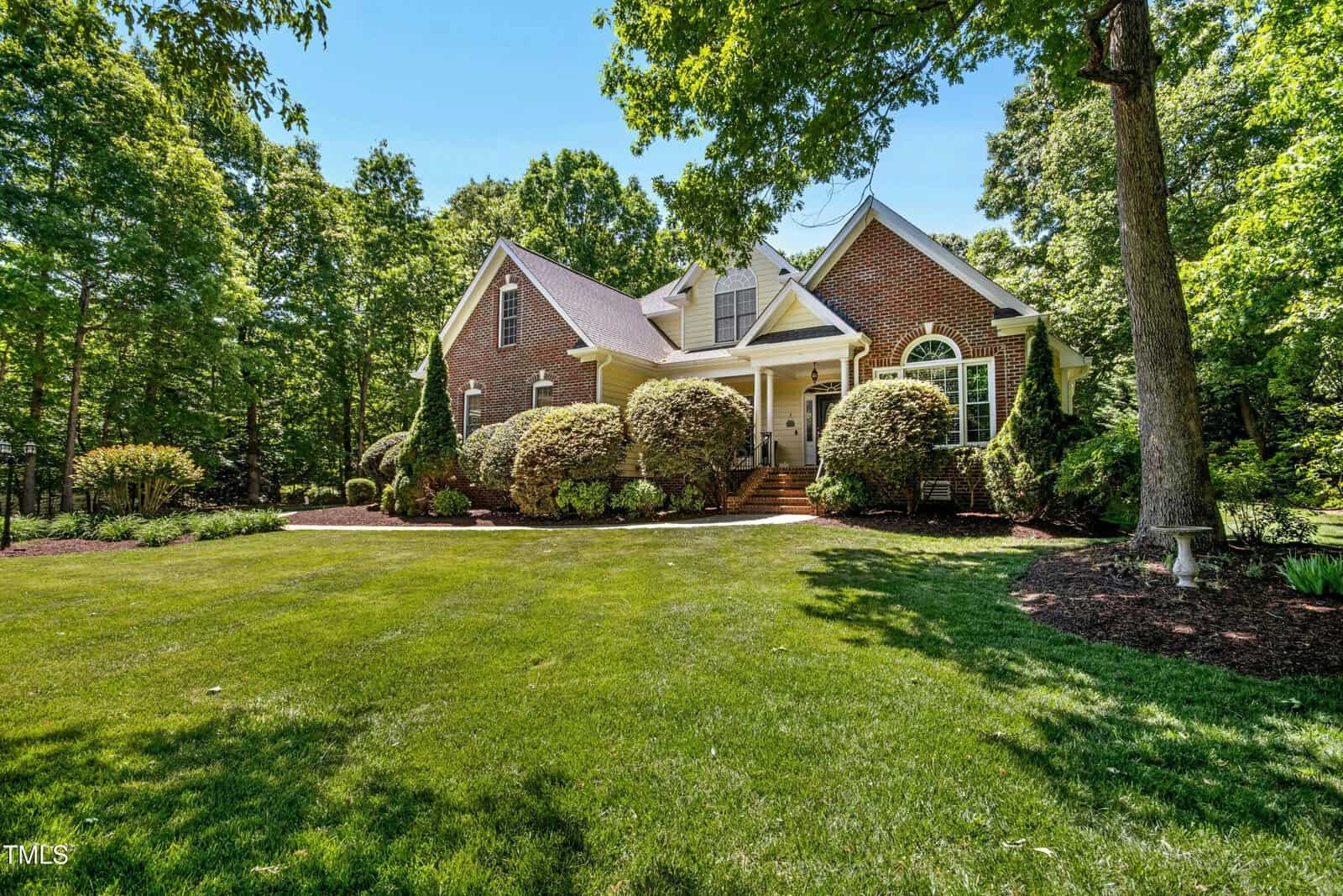 A brick house with a steep roof, white trim, and a covered front porch sits behind neatly trimmed bushes and a large green lawn, surrounded by tall trees and landscaped gardens on a sunny day.