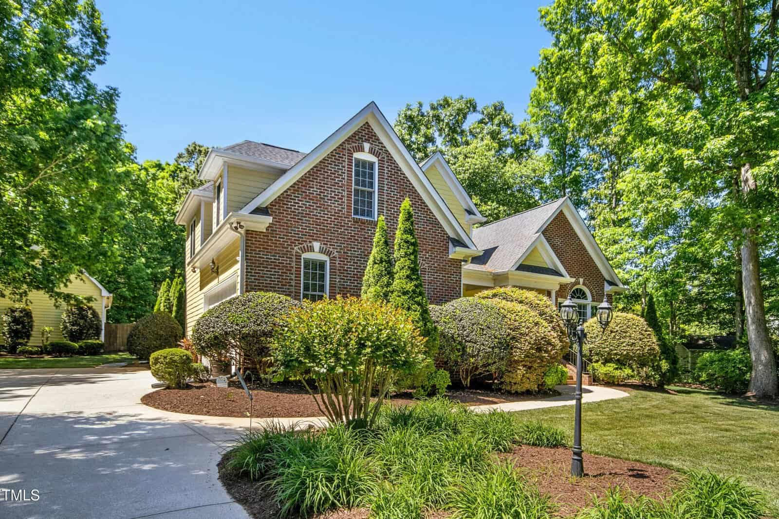 A two-story suburban house with red brick and beige siding, surrounded by well-manicured bushes, trees, and a curved driveway, under a clear blue sky.