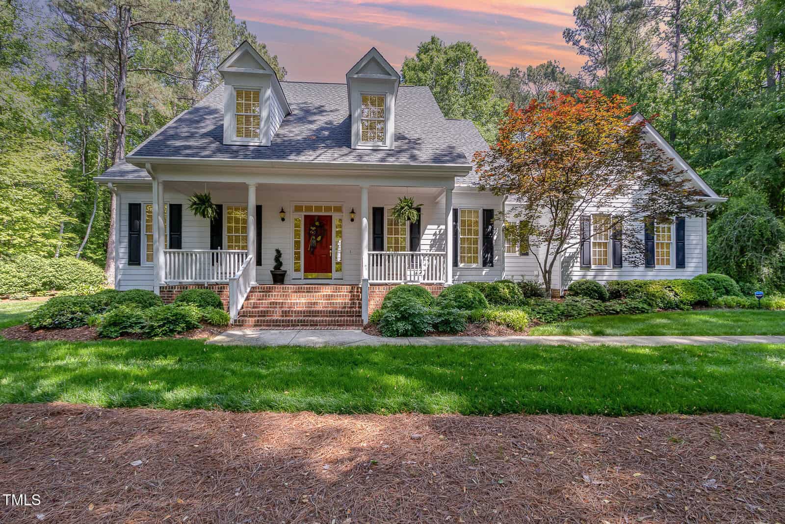 A charming white house with a large front porch, black shutters, two dormer windows, and a red front door, surrounded by green lawns, shrubs, and tall trees under a colorful sky.